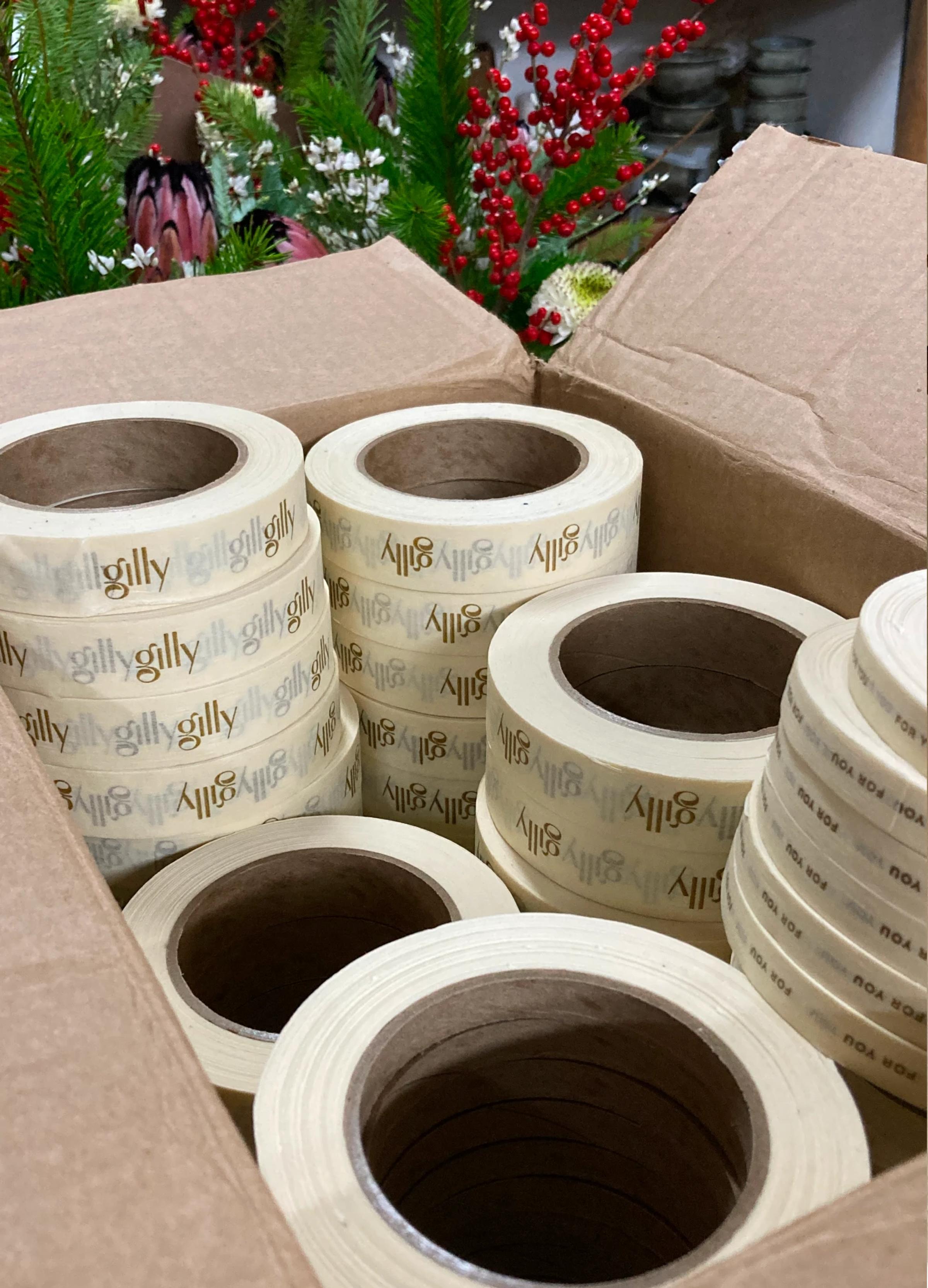 A box filled with rolls of decorative tape printed with "gilly," surrounded by colorful floral arrangements.