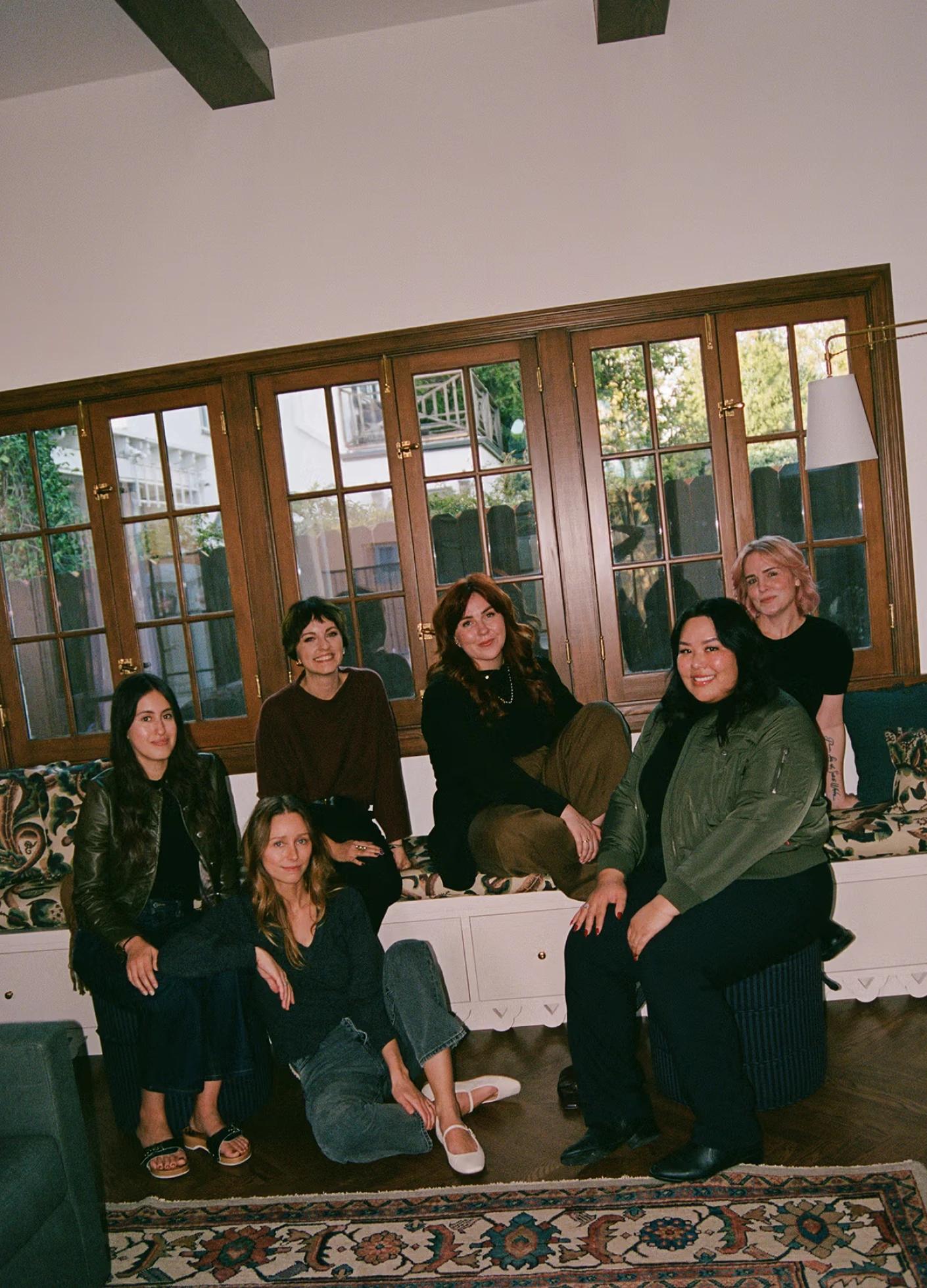Six women sit together on a couch, smiling and posing in a cozy, well-lit room with large windows.