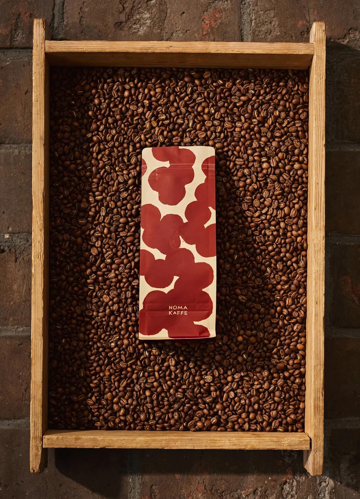 A coffee bag with a red abstract design sits atop a wooden tray filled with roasted coffee beans.