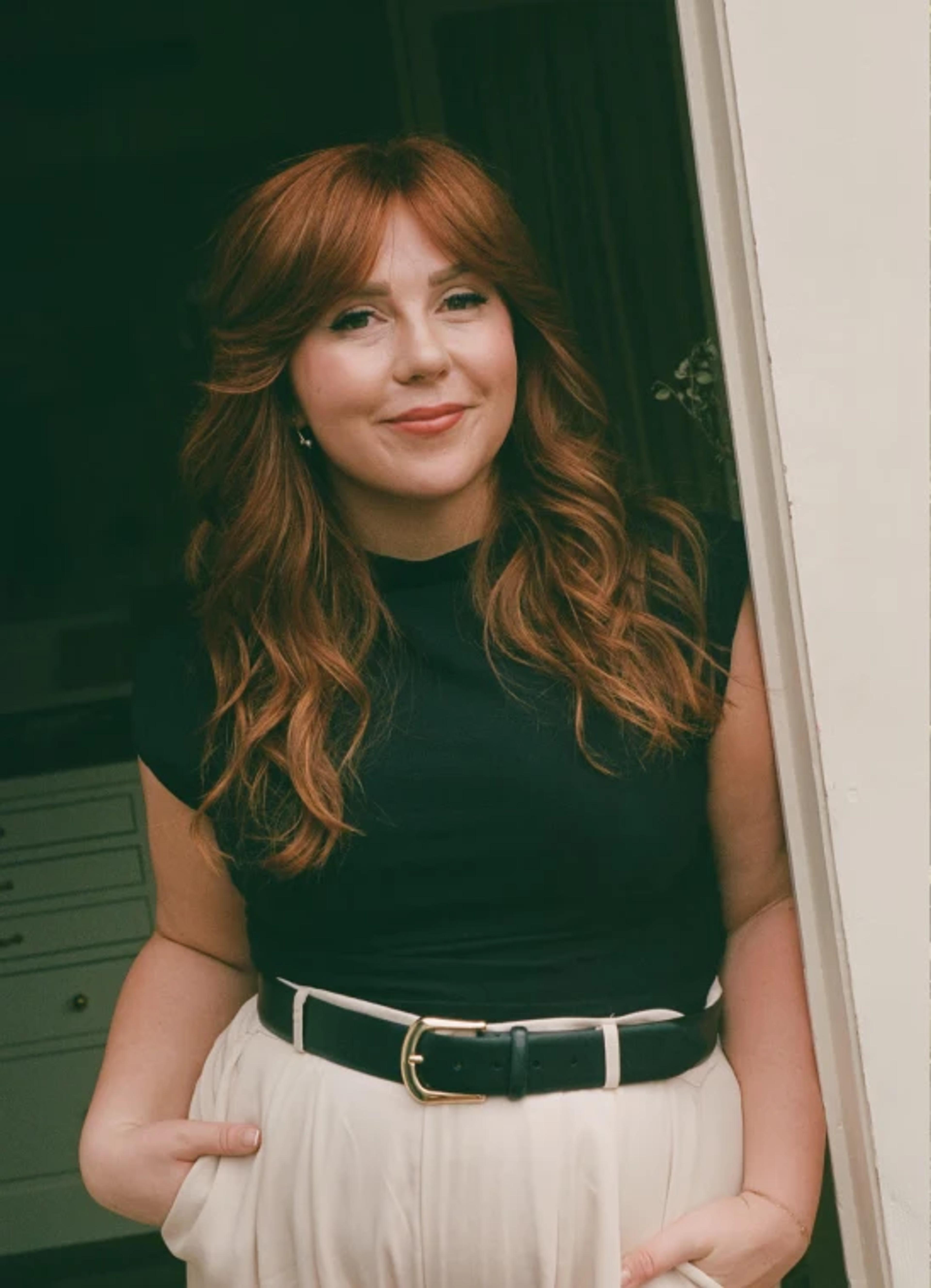 A woman with long, wavy red hair smiles while leaning against a doorway, wearing a black top and high-waisted white pants.