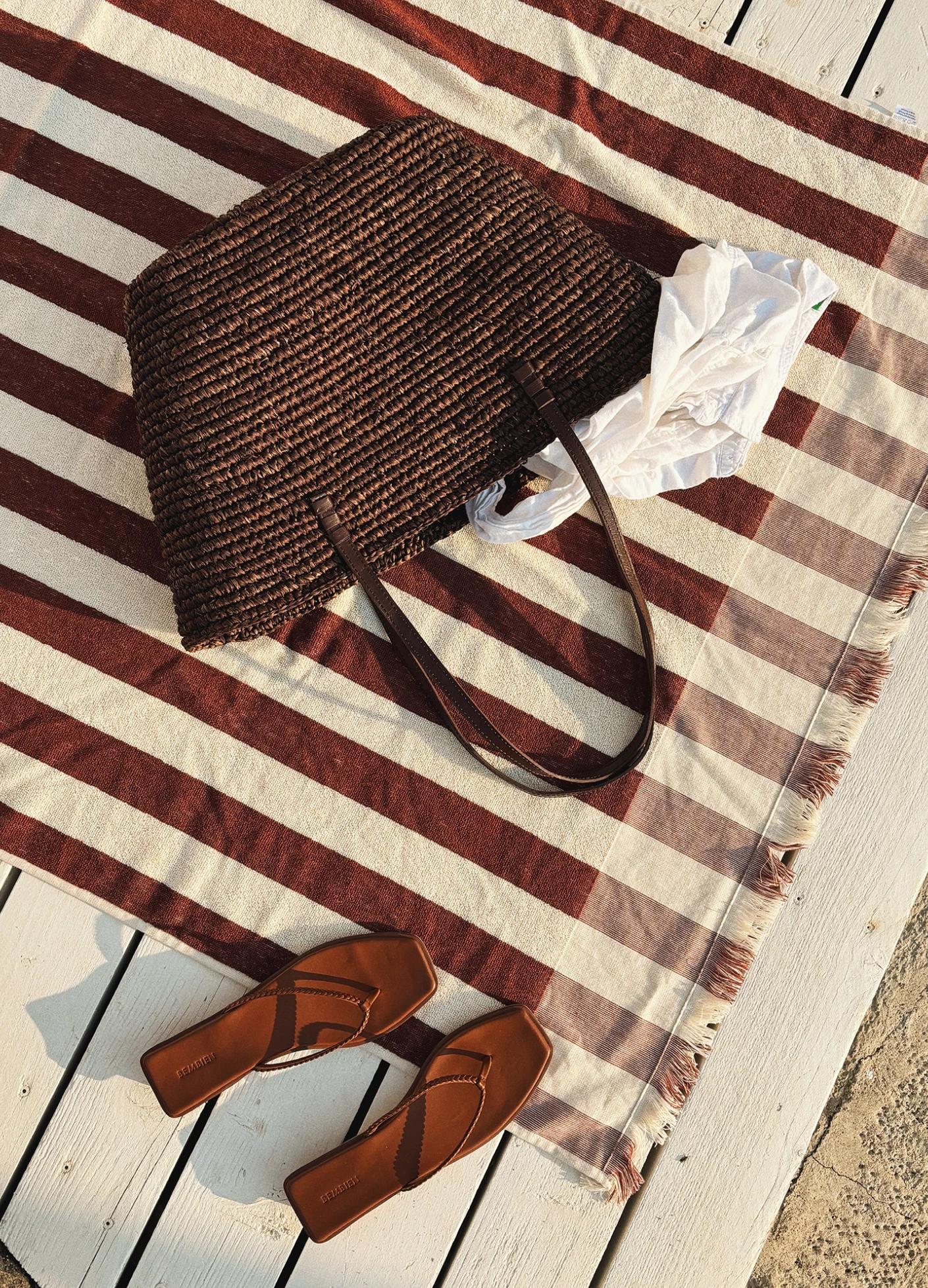 A woven brown tote bag and brown flip-flops resting on a striped beach towel laid on a wooden surface.
