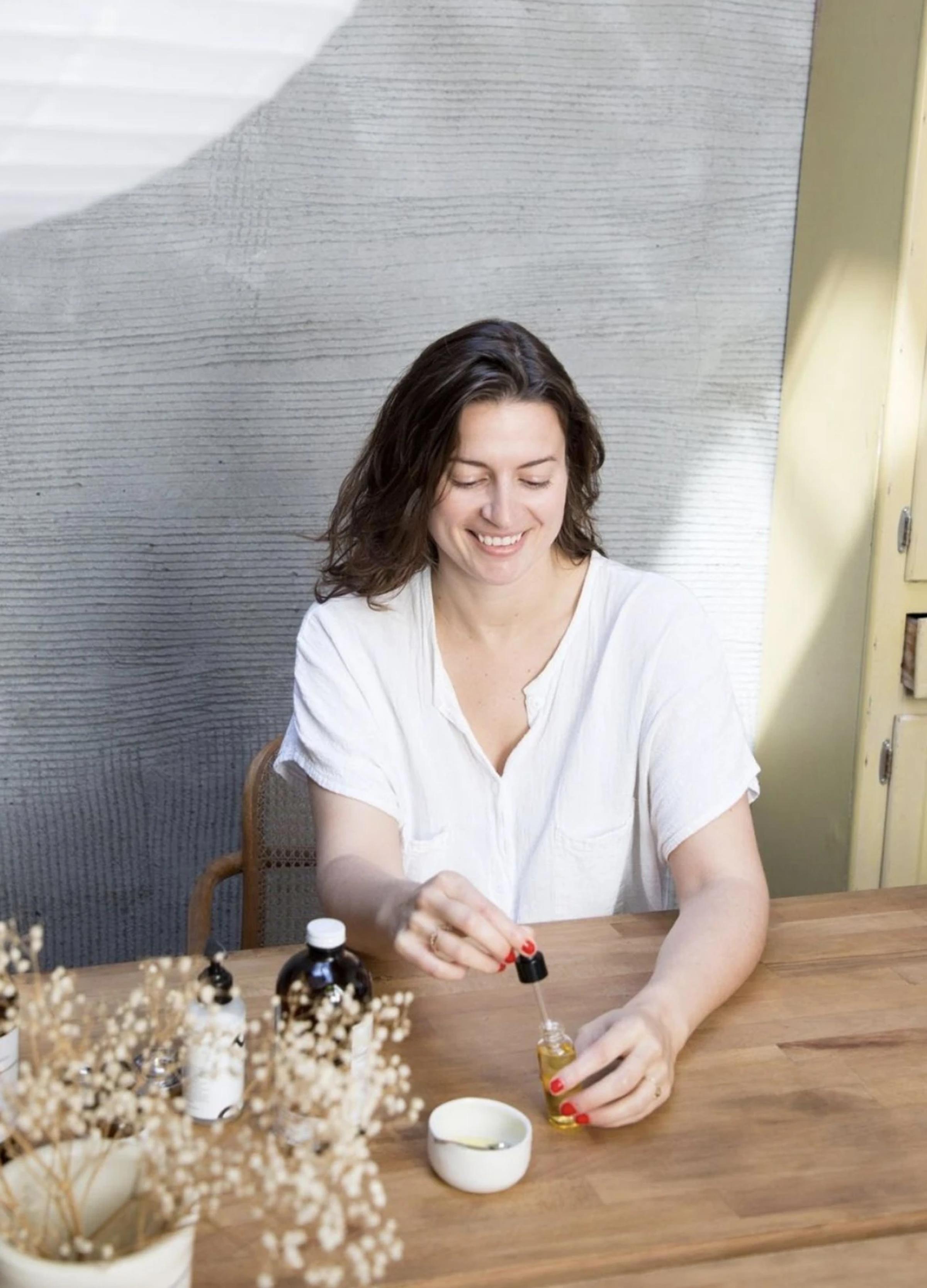 A woman with wavy hair smiles while using a dropper to dispense oil into a small dish on a wooden table.