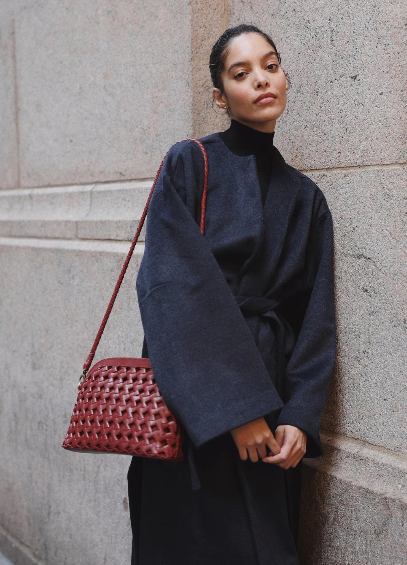 A woman in a dark oversized coat stands against a textured wall, holding a woven red handbag.