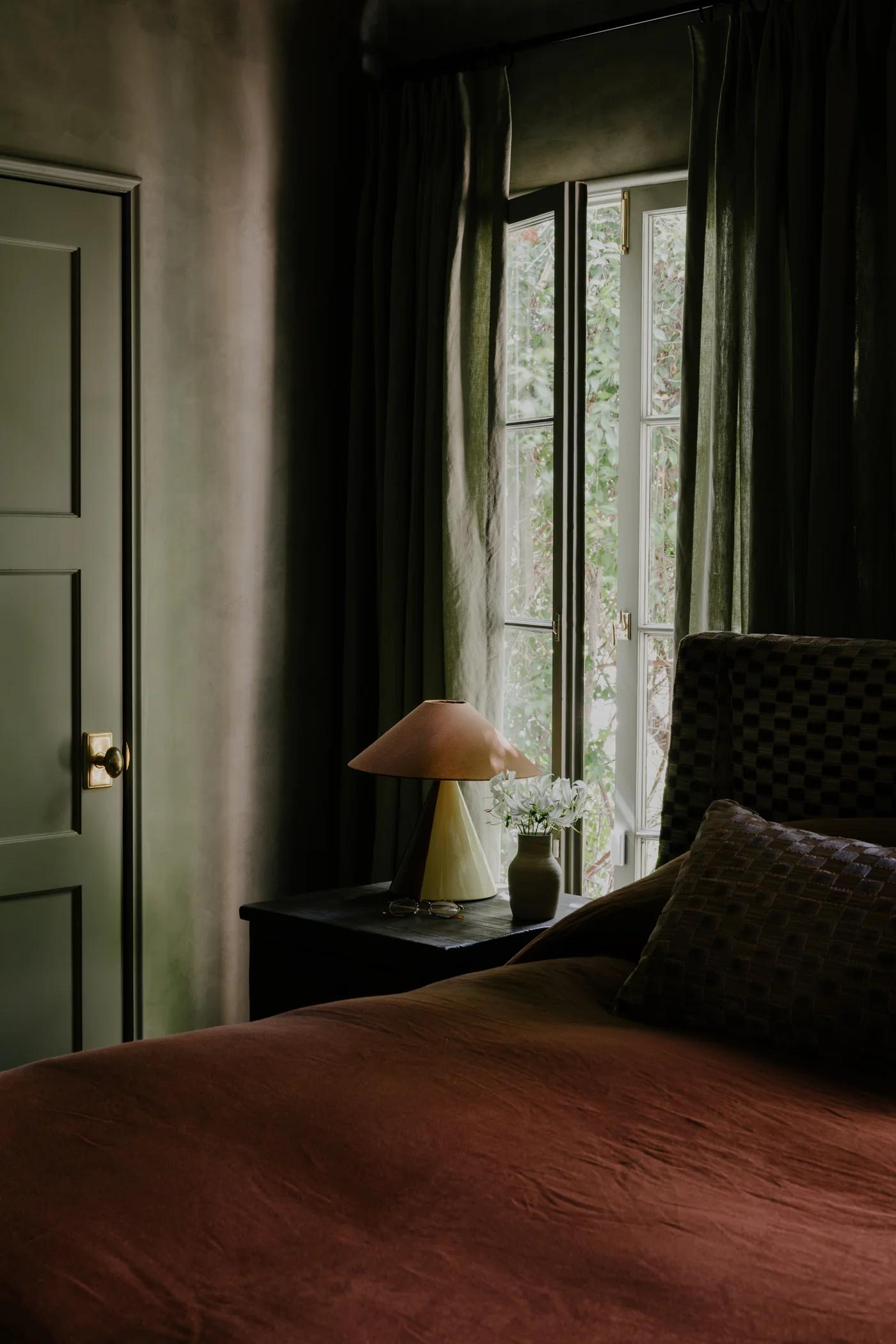 Cozy bedroom with a brown bedspread, a stylish lamp, a vase of flowers, and sunlight streaming in through green curtains.