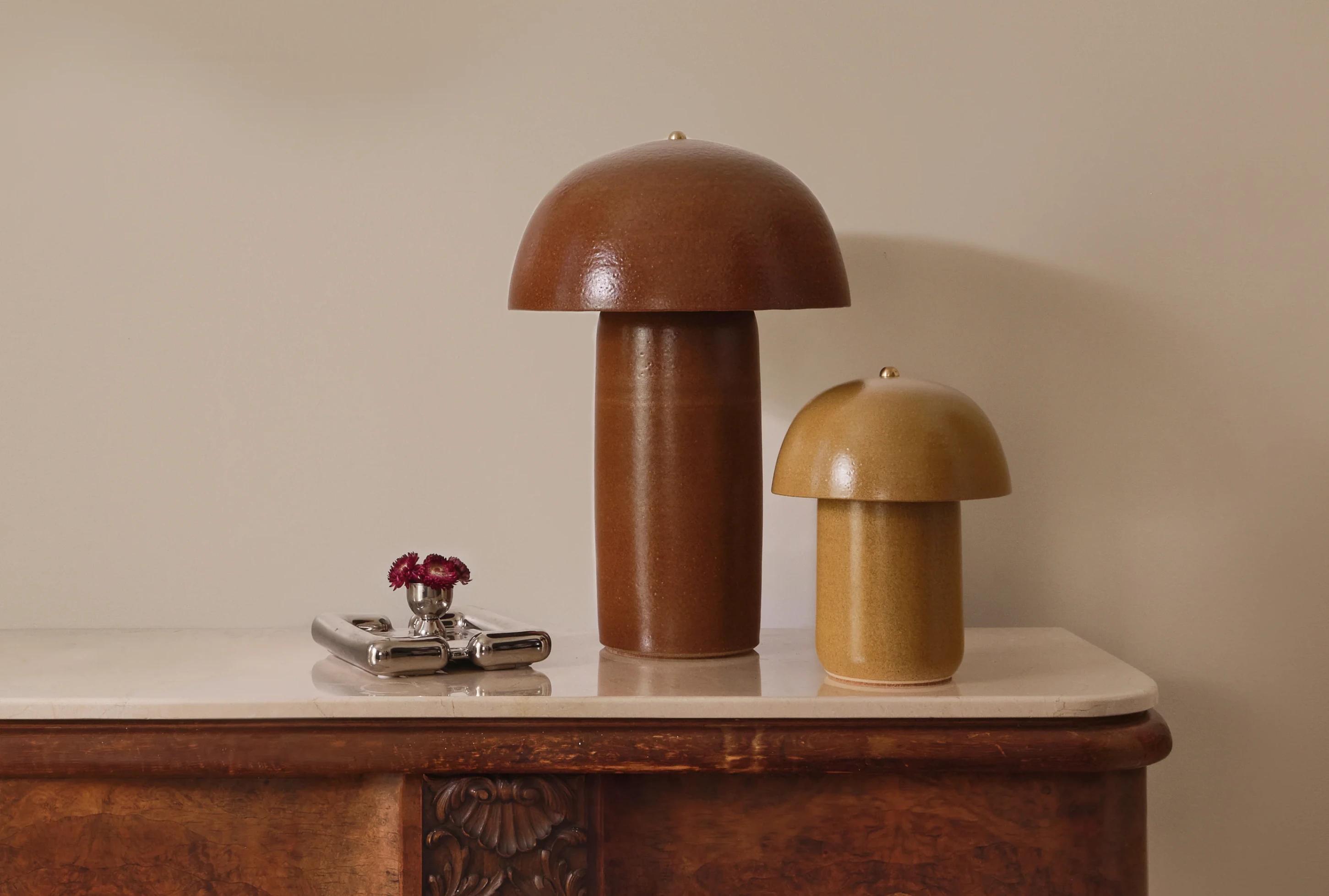 Two ceramic mushroom-shaped lamps in brown and beige, alongside a silver planter with small purple flowers on a wooden table.
