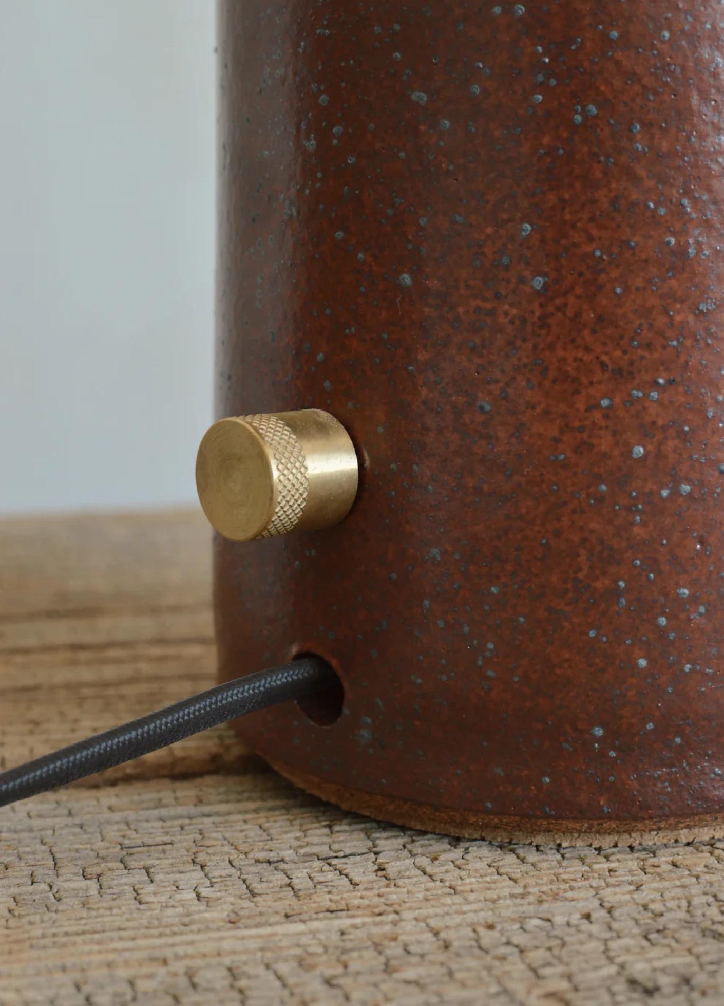 A textured brown ceramic lamp base with a brass knob and a black cord on a wooden surface.