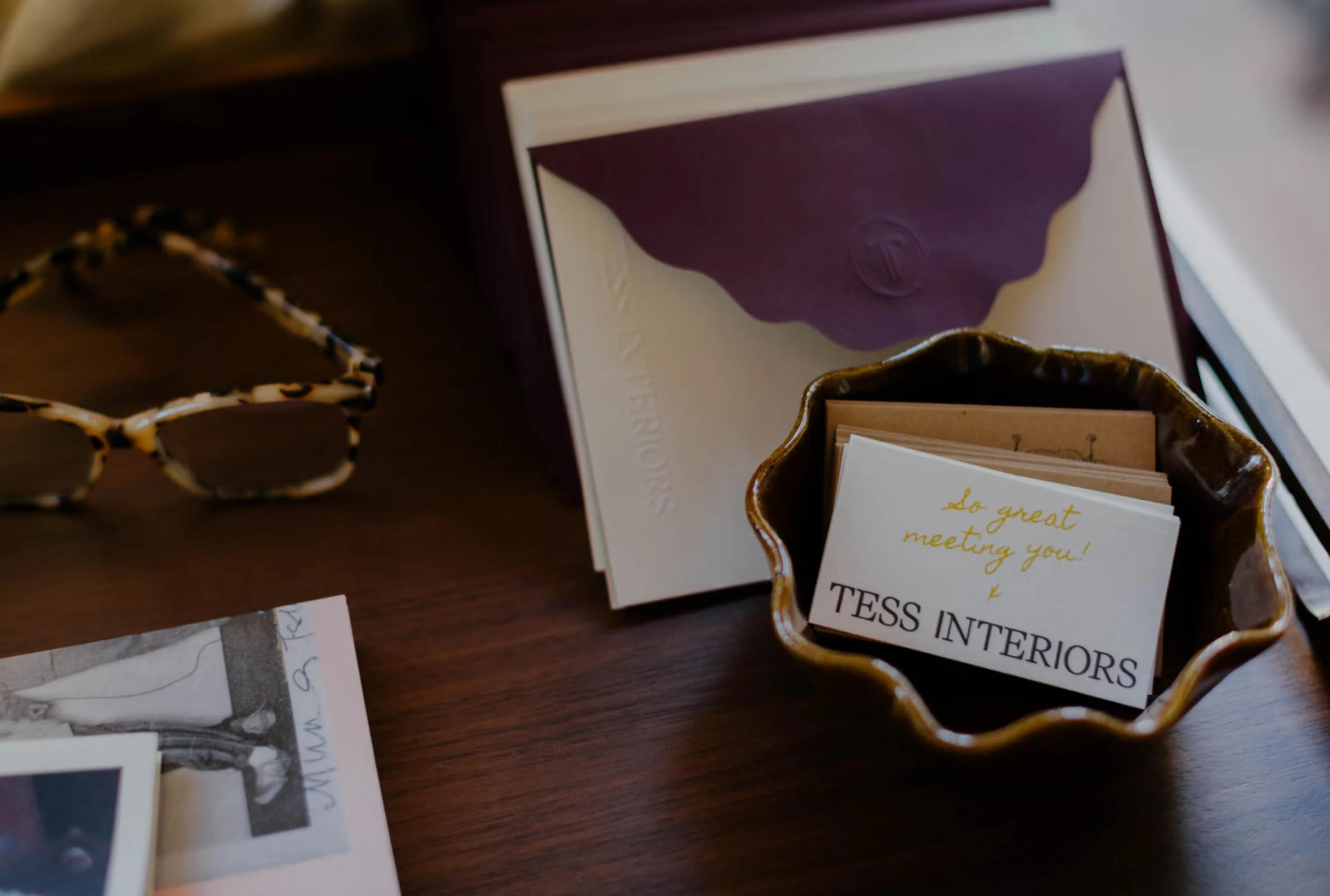 A small bowl holds business cards with "TESS INTERIORS," beside a pair of patterned glasses and a purple envelope.