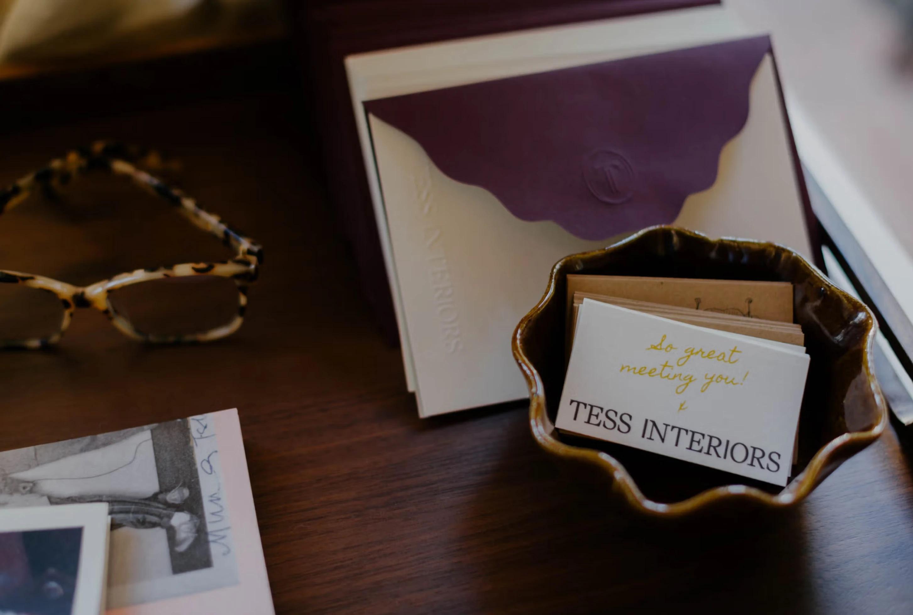 A small bowl holds business cards with "TESS INTERIORS," beside a pair of patterned glasses and a purple envelope.