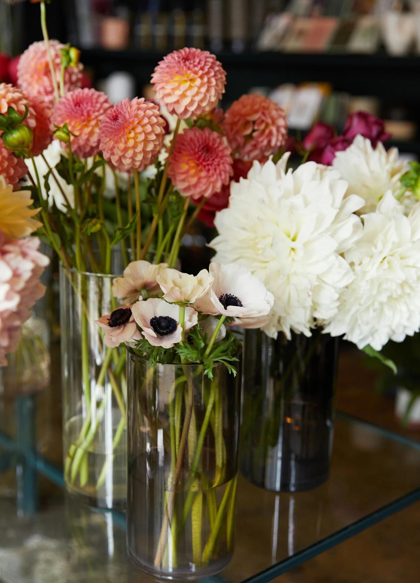 A variety of colorful flowers, including pink dahlias and white peonies, arranged in clear vases atop a glass table.