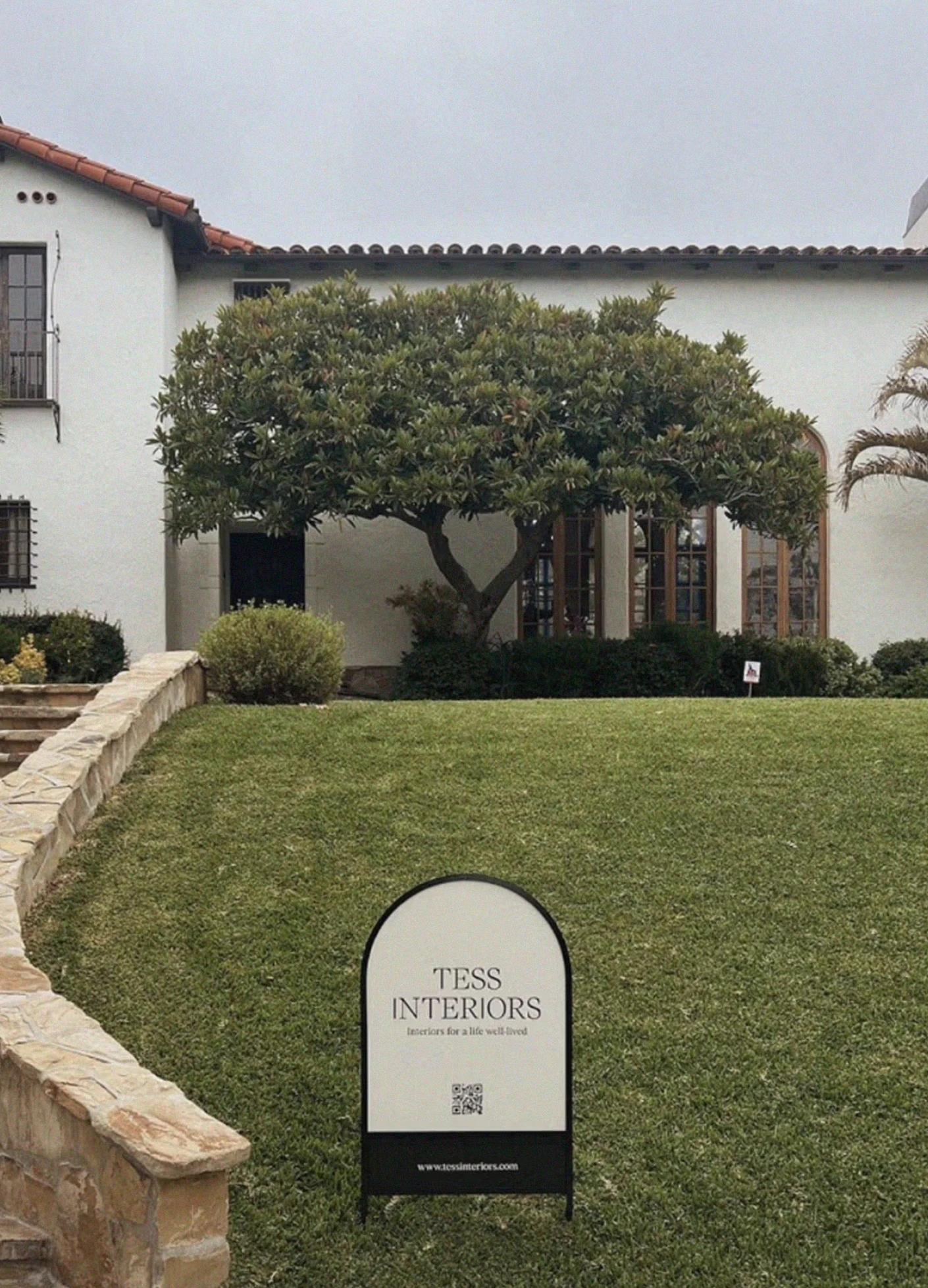 A well-maintained lawn with a large tree, a stone path, and a sign for "Tess Interiors" in front of a white house.