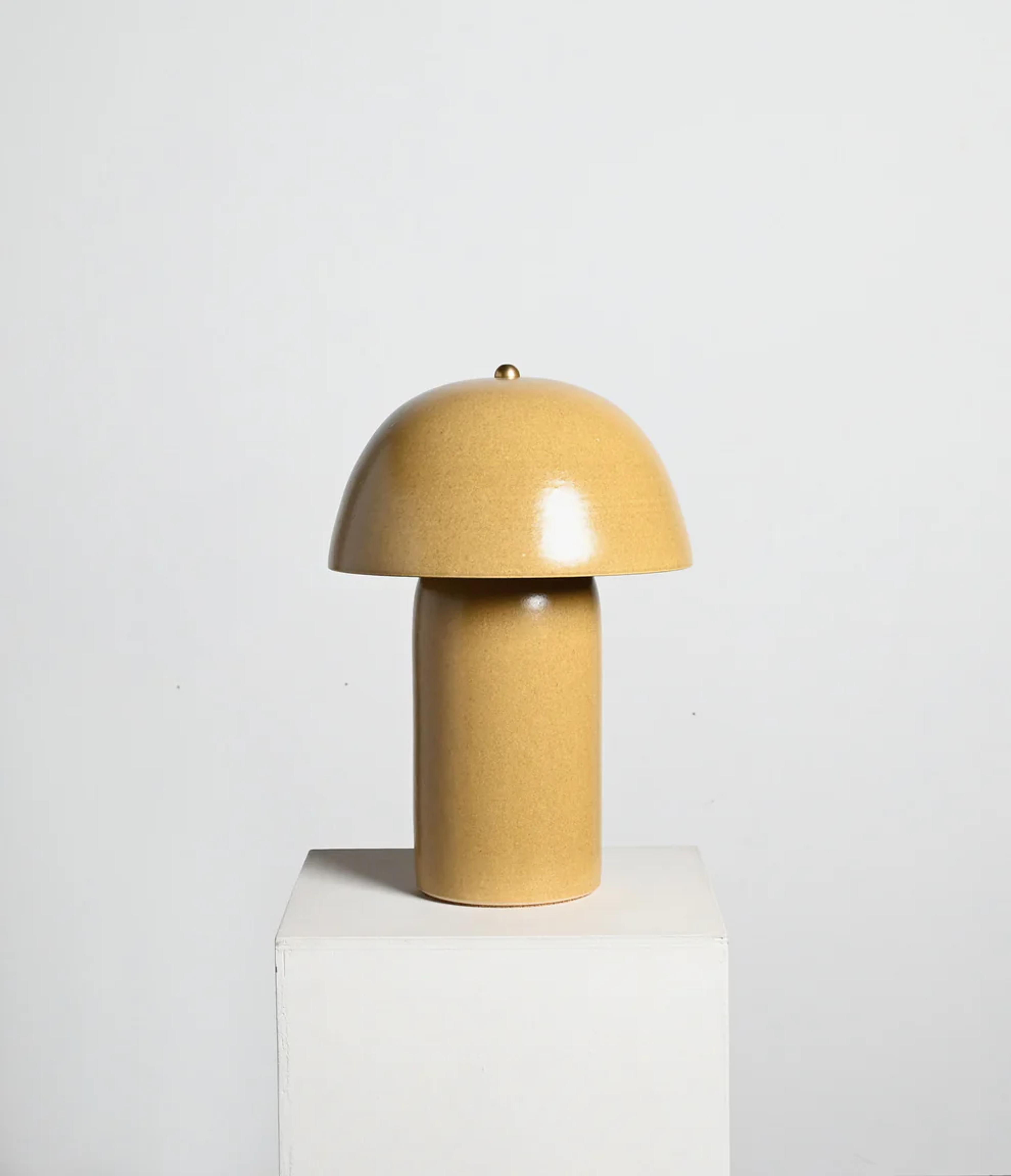 A modern, sculptural lamp with a rounded dome shade and a cylindrical base in a warm beige tone, placed on a white pedestal.