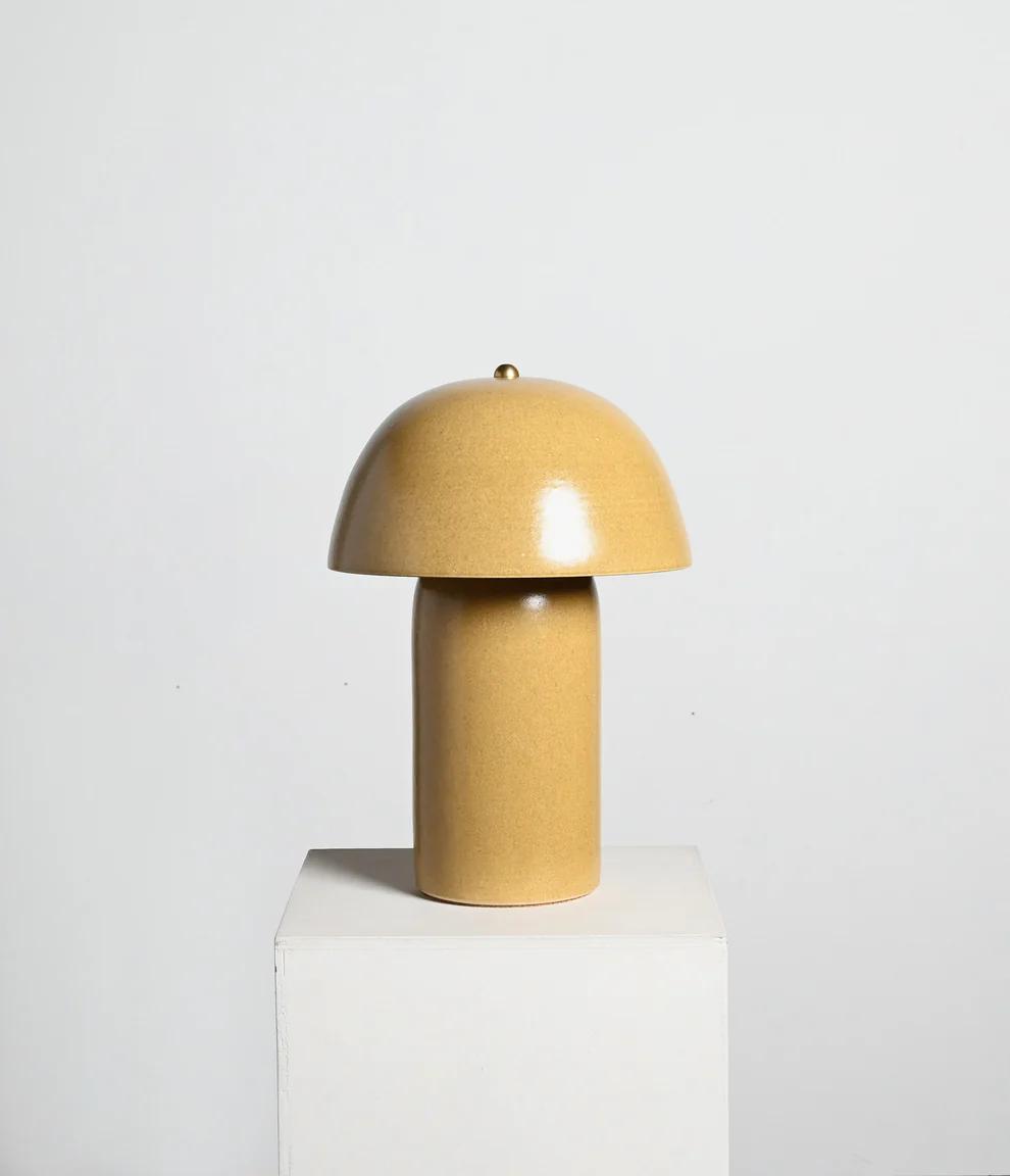 A modern, sculptural lamp with a rounded dome shade and a cylindrical base in a warm beige tone, placed on a white pedestal.