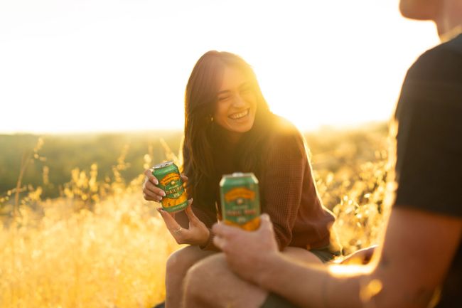 A woman holds a can of non-alcoholic Trail Pass IPA while chatting with a friend in an outdoor setting.