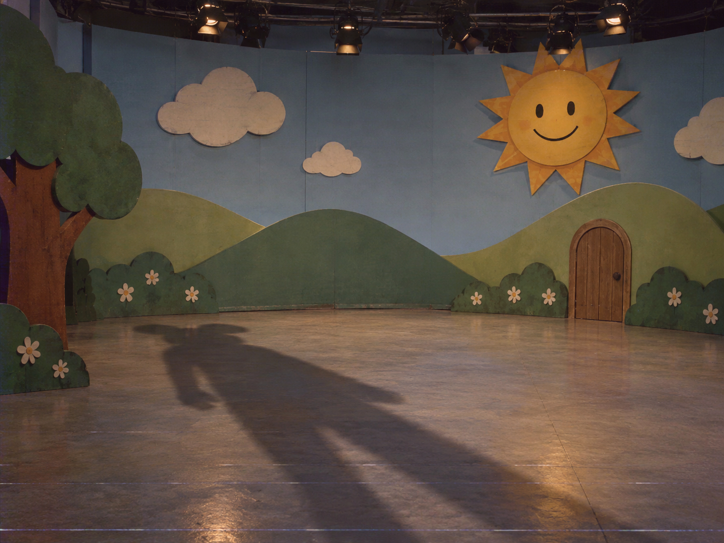 Großer maskottchenartiger Schatten auf einem leeren Kindershow-Set mit lächelnder Sonne und gemalten Hügeln