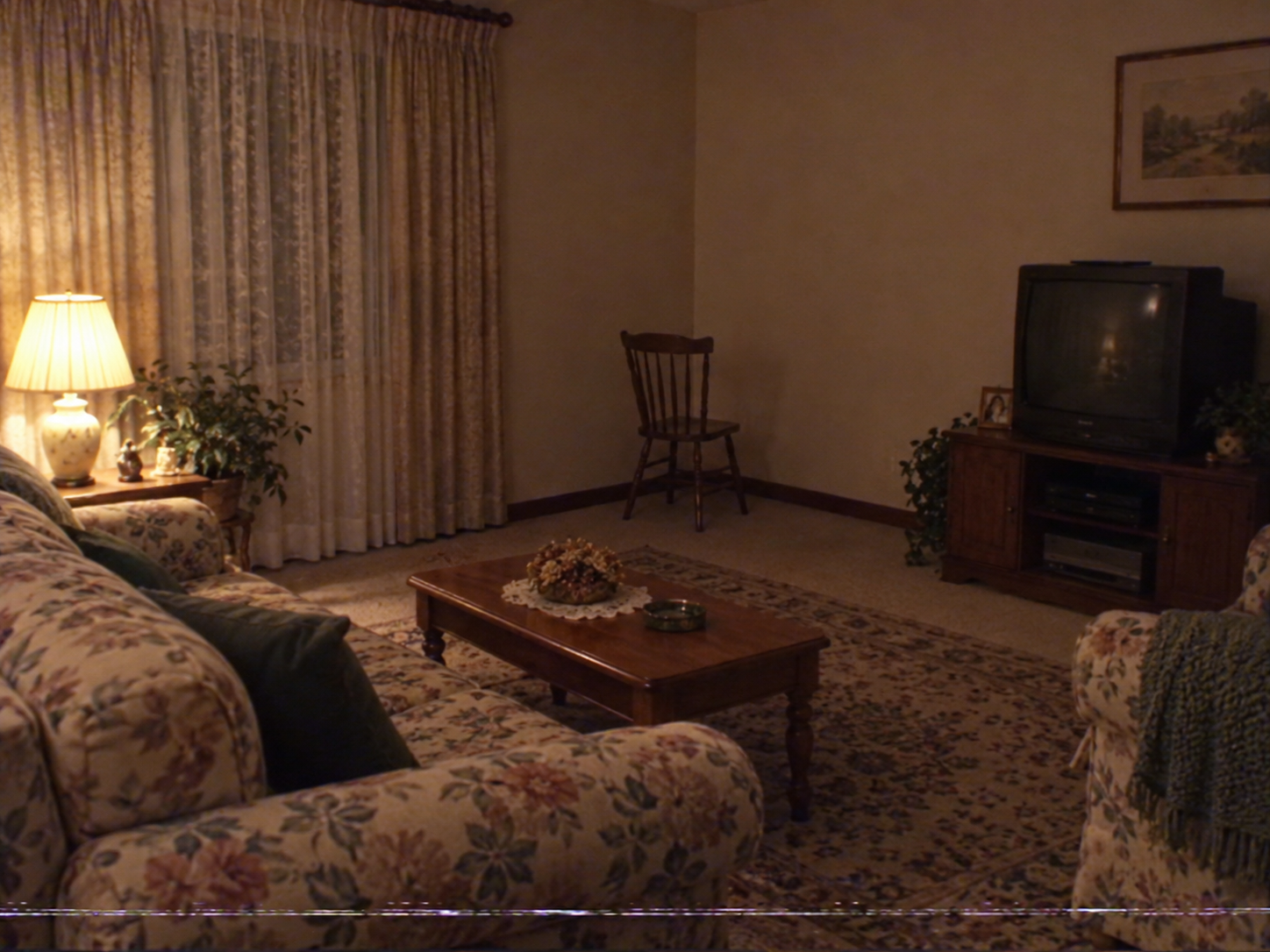 Wohnzimmer mit Röhrenfernseher, gemustertem Sofa und Sessel sowie einem Holzstuhl nahe der Zimmerecke