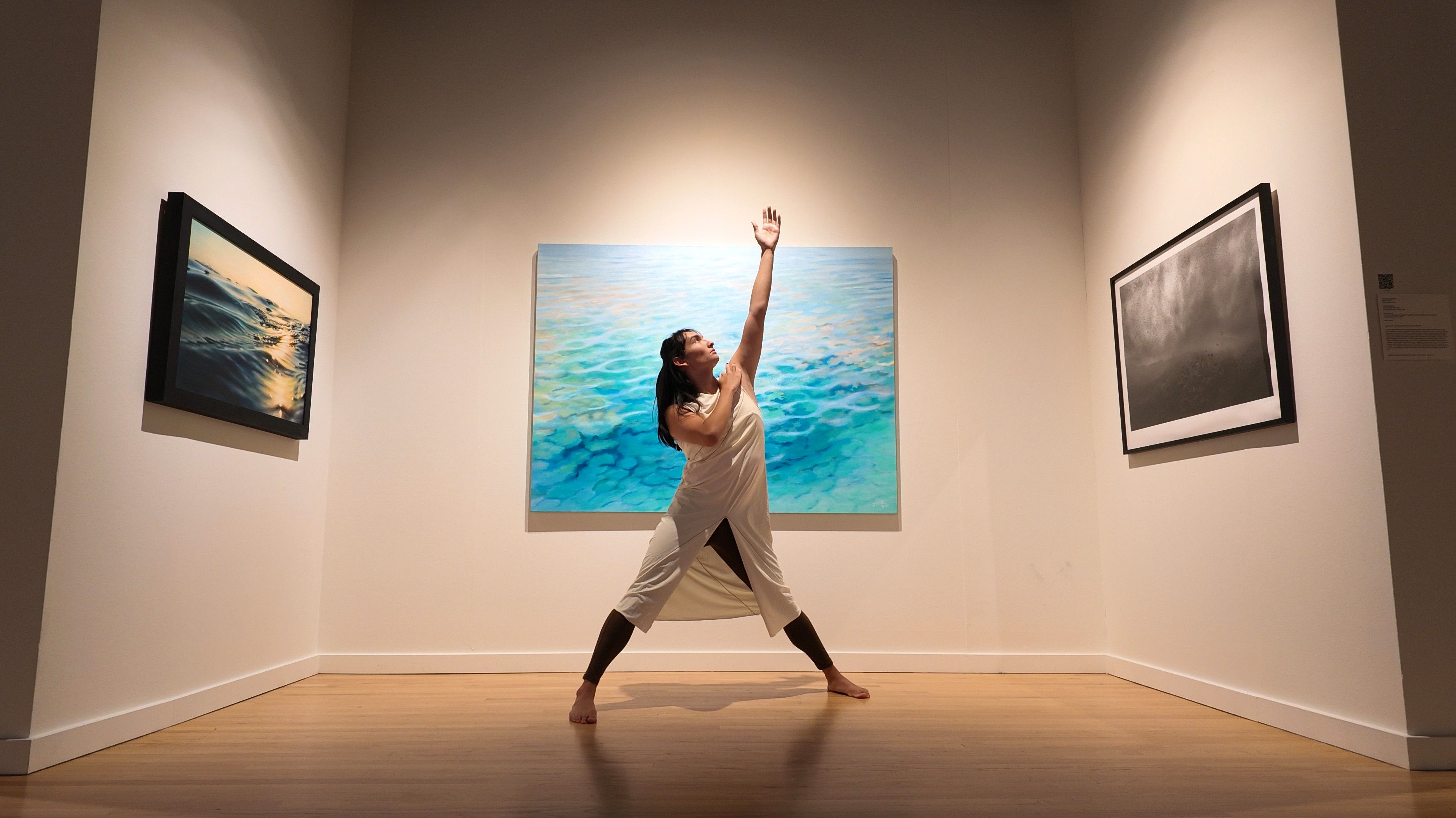 Dancer posing with one arm up in front of a painting of a seascape.