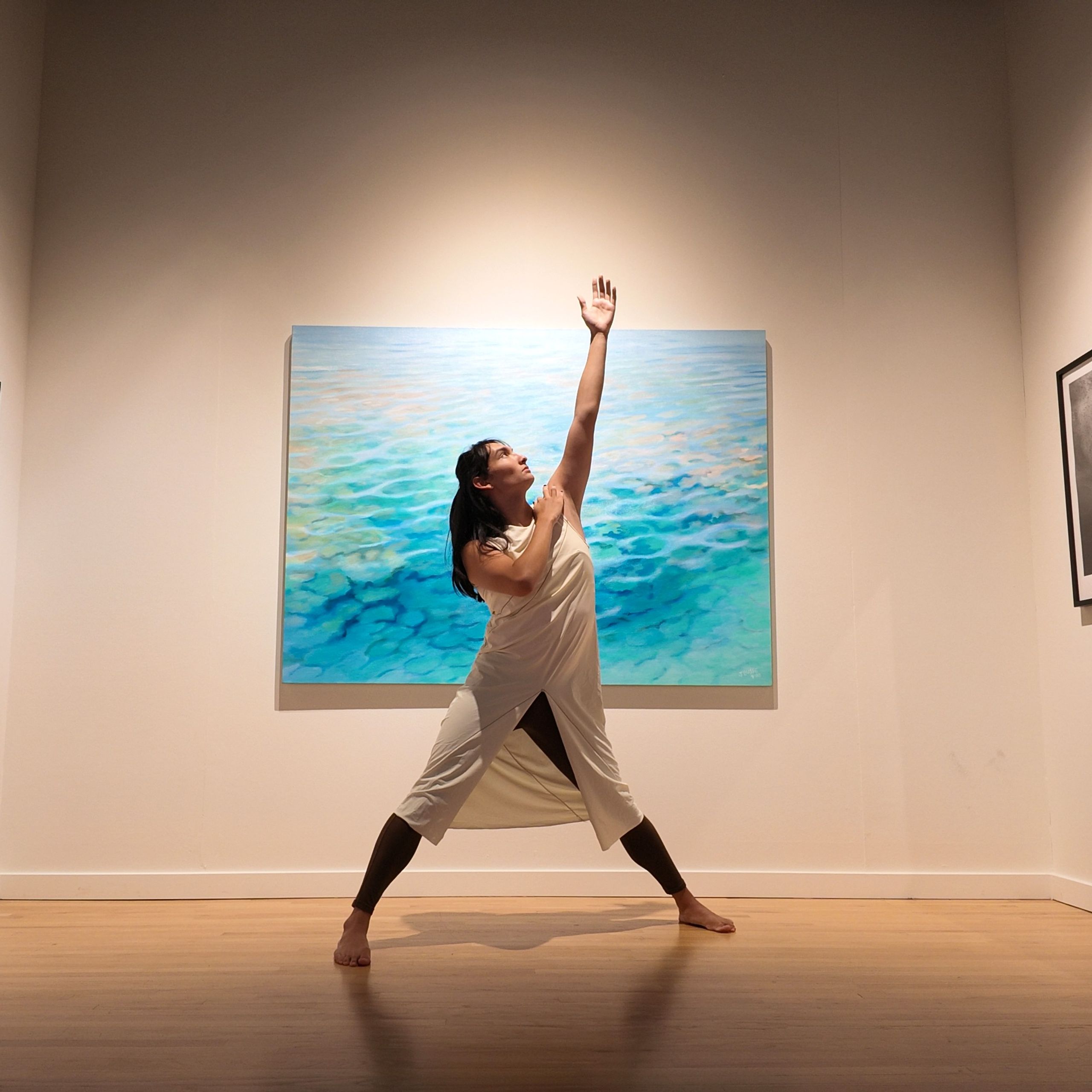 Dancer posing with one arm up in front of a painting of a seascape.