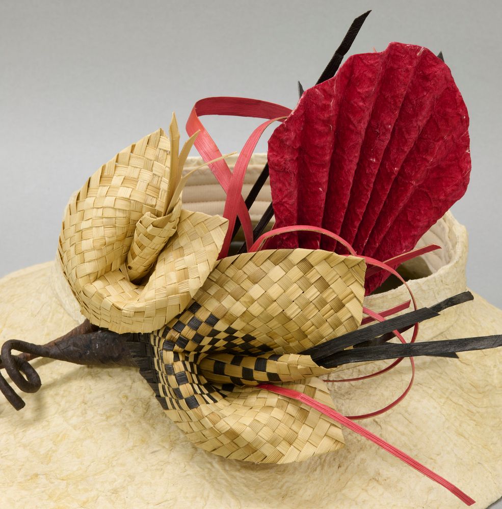 Closeup of open-topped hat made with light brown kapa, decorated with woven flowers.