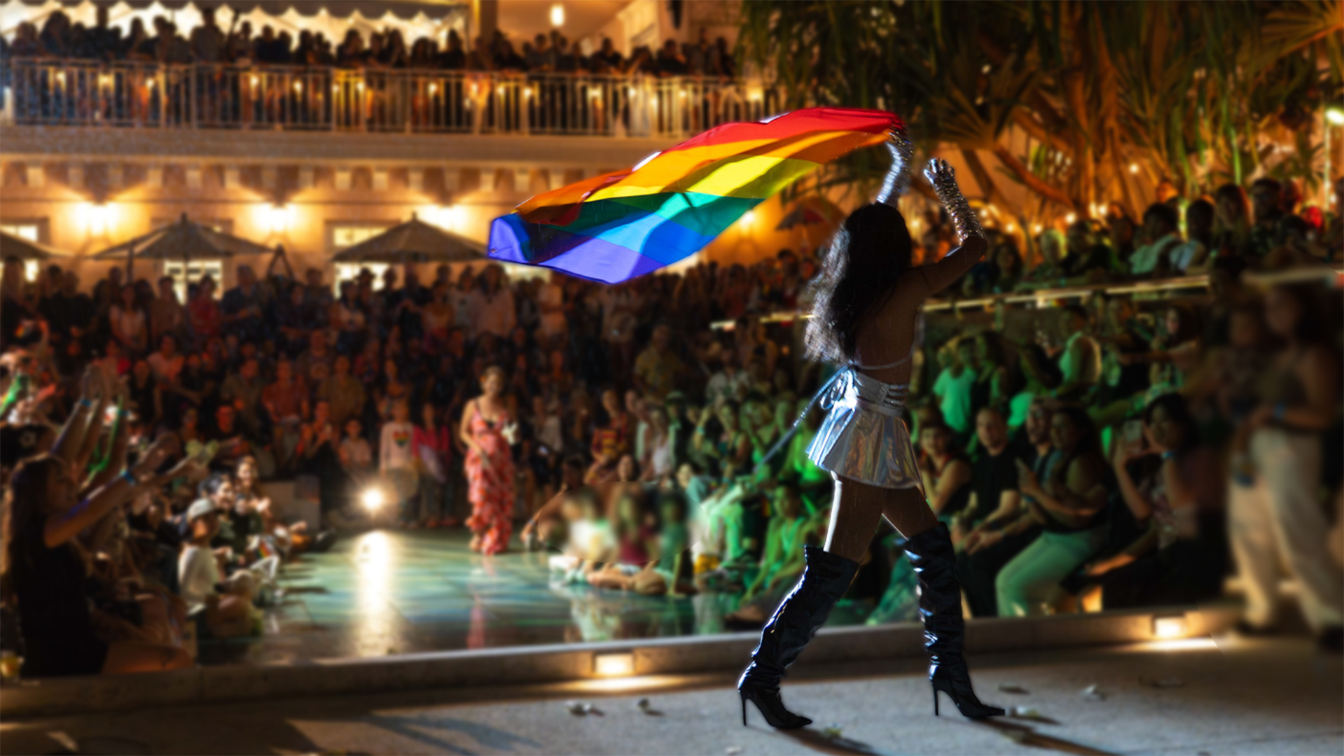 Evening in an outdoor space with colorful lighting and performer holding a rainbow flag.