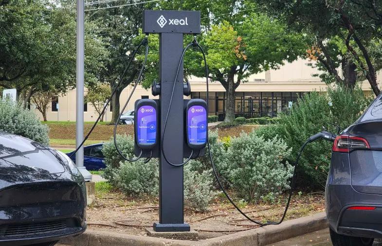 two electric cars are being charged at a xeal charging station in a parking lot .