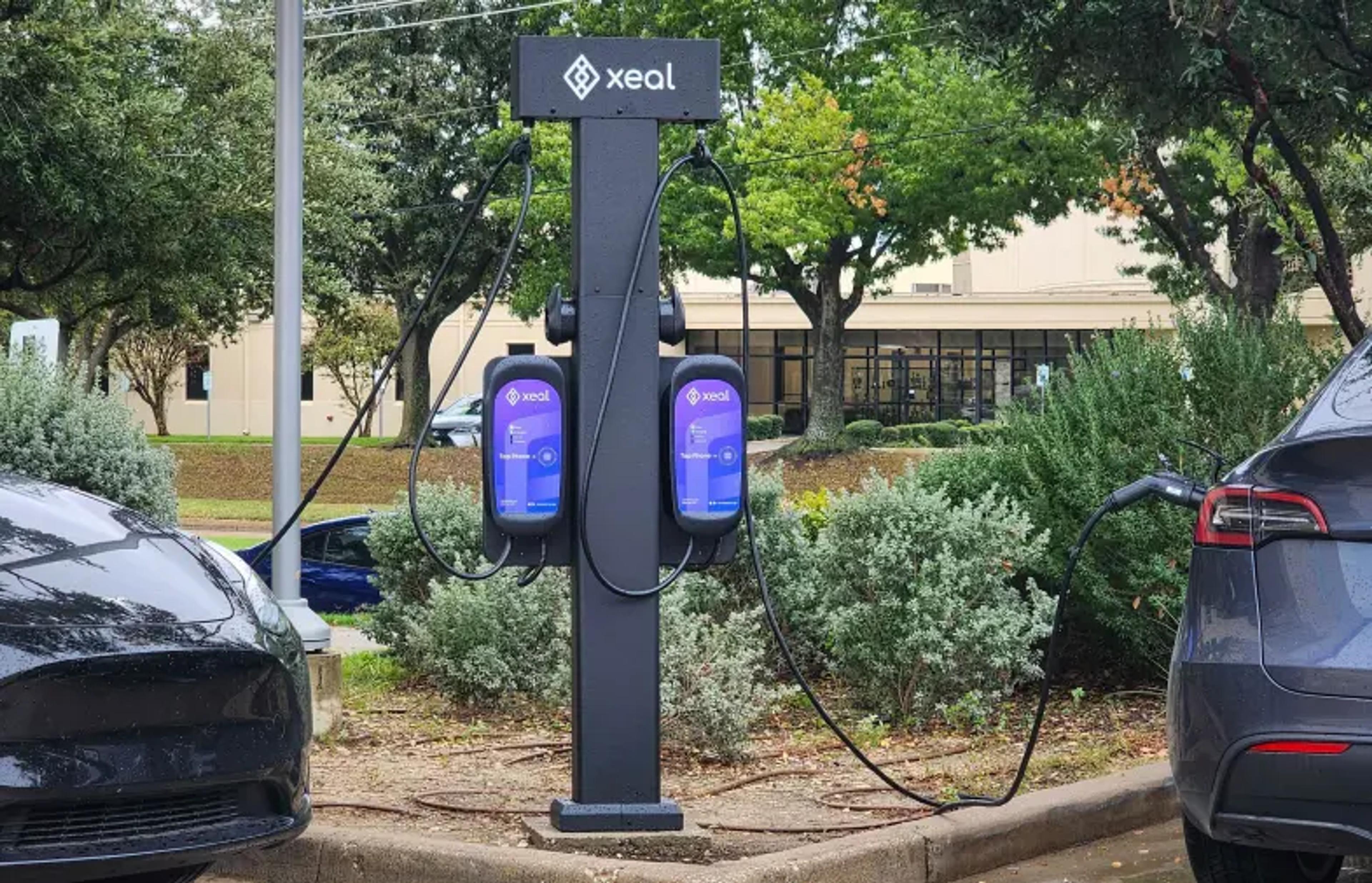two electric cars are being charged at a xeal charging station in a parking lot .