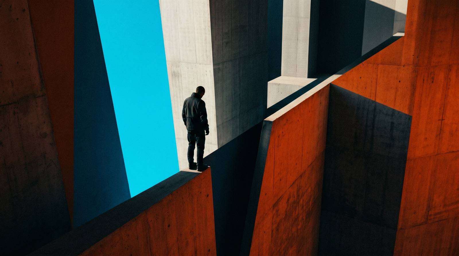 A small figure standing at the brink of a sharp drop in bold geometric architecture, deep shadow one side.