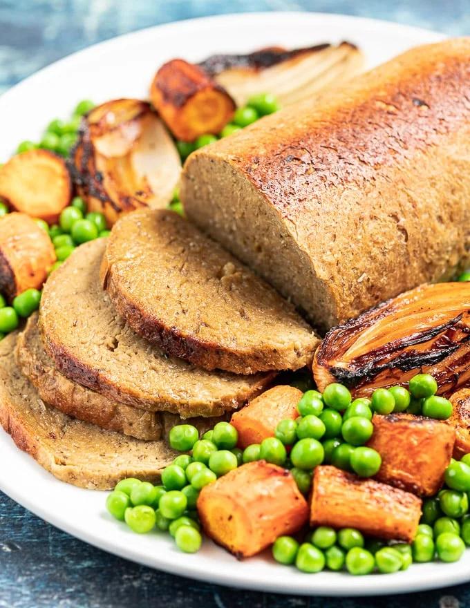 A sliced vegan roast on a plate with peas, carrots, and other veggies.