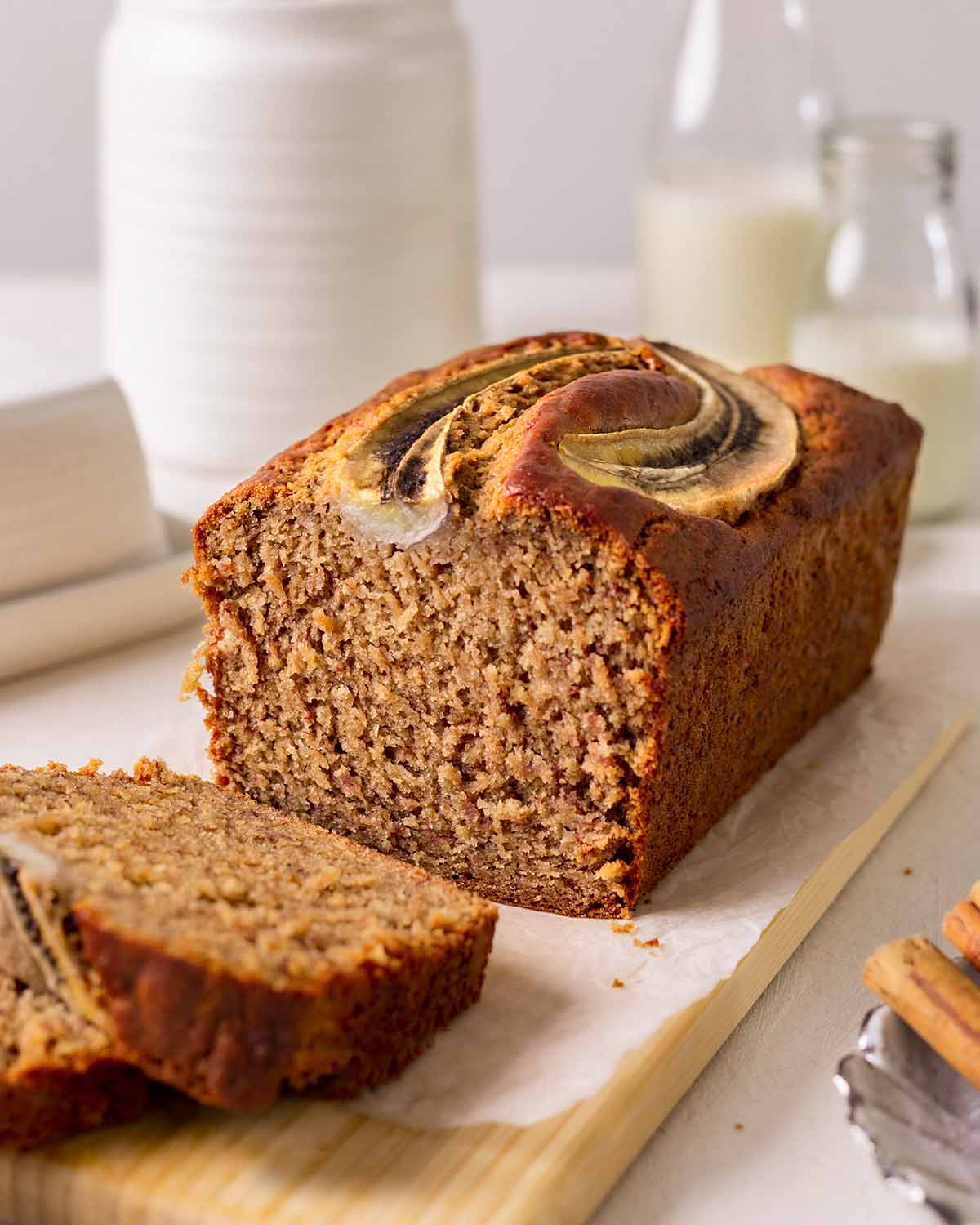Vegan banana bread on a serving board, sliced, with jars of plant milk in the background. 