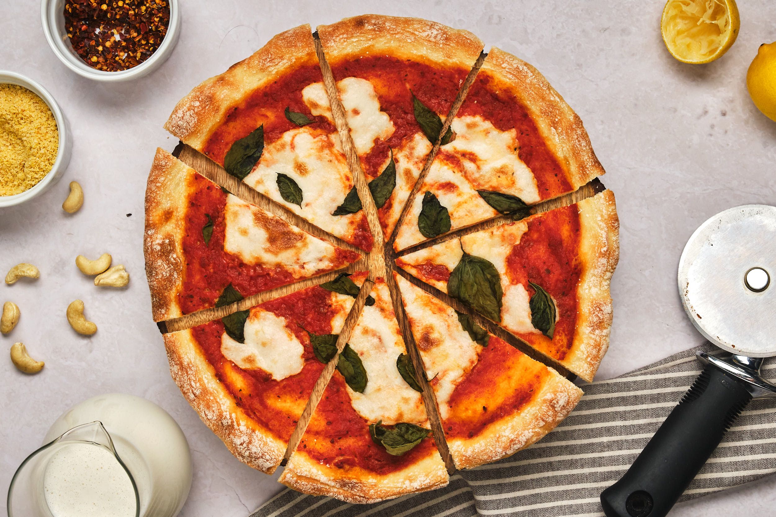 A sliced vegan pizza with a jar of liquid mozzarella to the side, as well as cashews, vegan parmesan, crushed red pepper, lemons, and a pizza cutter.