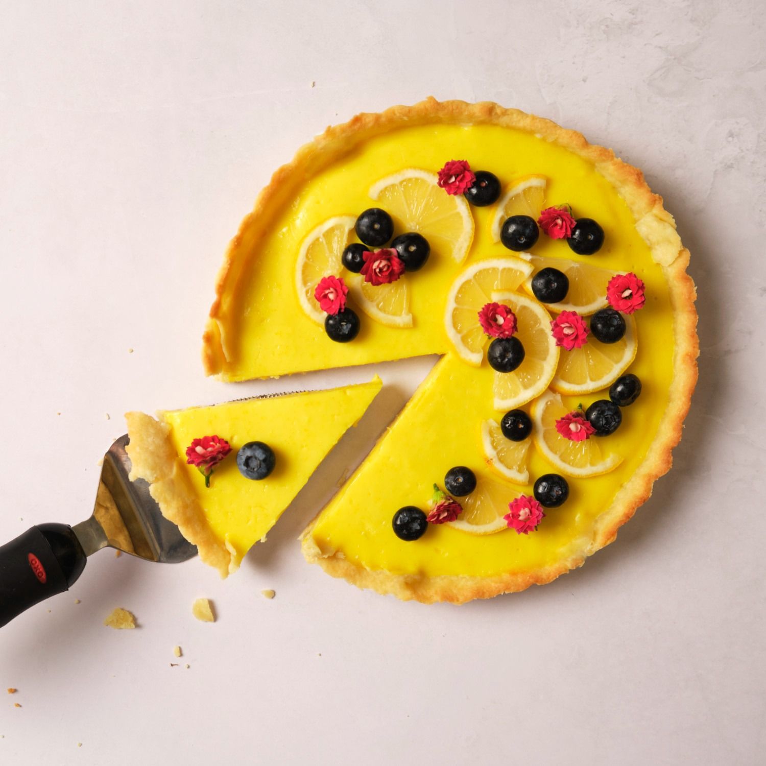 A vegan lemon tart topped with berries, flours, and lemon slices, with one slice being pulled out with a pie server.