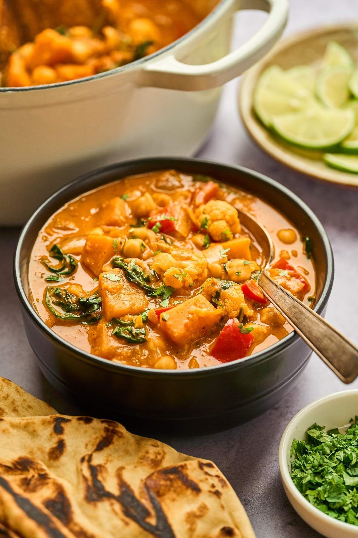 Vegetable Curry in a pot and in a bowl next to flatbread and small bowls with lime slices and chopped parsley.