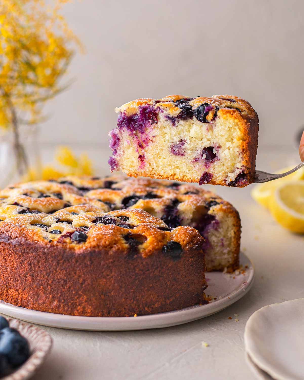 A slice of lemon blueberry cake on a cake server, just above the rest of the cake. 