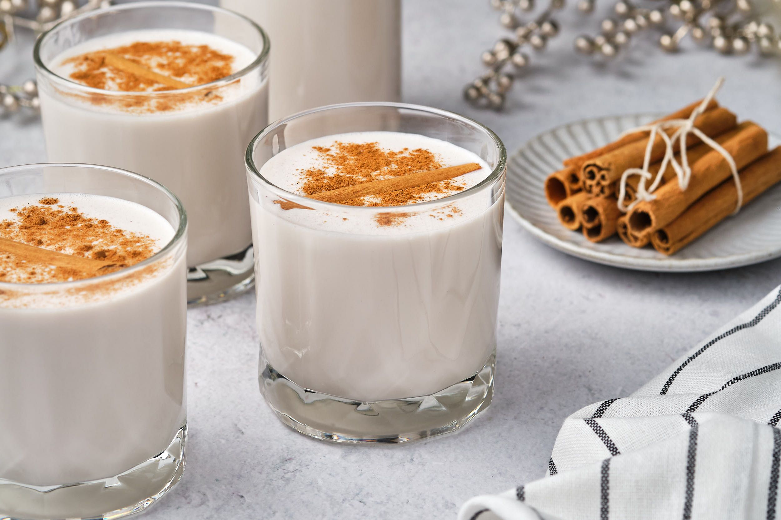 Three glasses of vegan coquito topped with cinnamon; cinnamon sticks and a bottle of coquito are in the background.