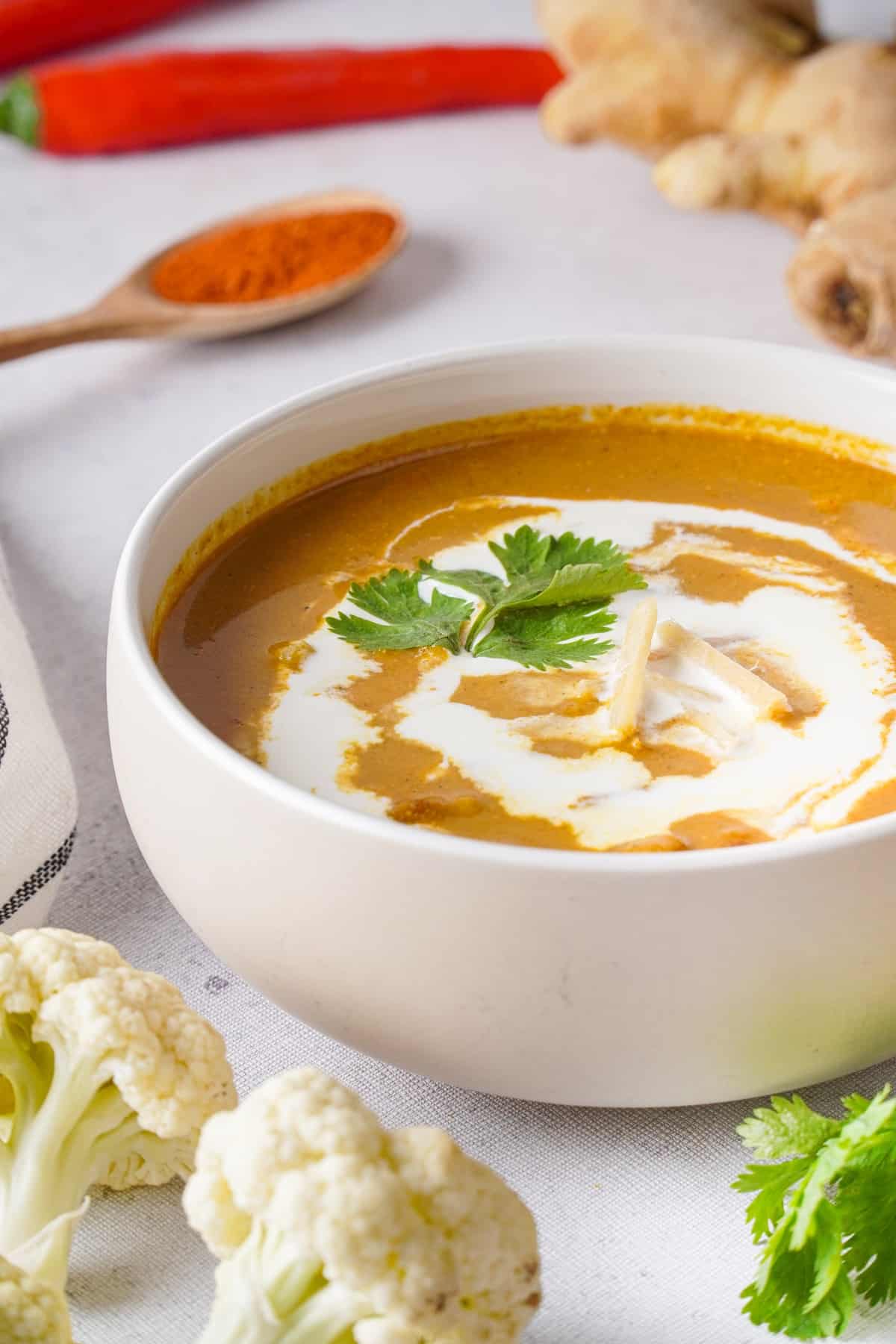 A bowl of cauliflower tikka masala soup surrounded by peppers, ginger, and cauliflower.