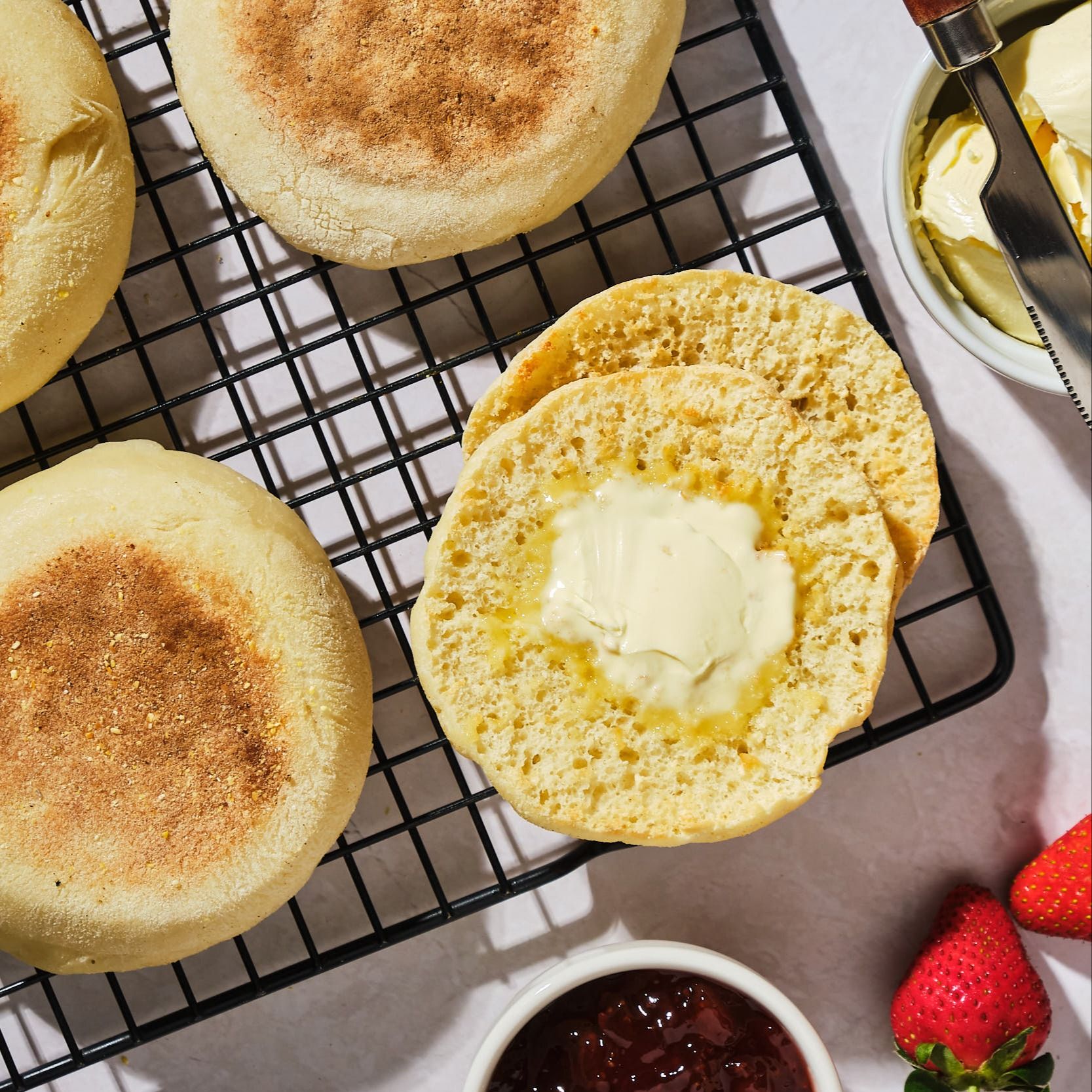 A top-down view of vegan English muffins on a wire cooling rack, with one muffin cut in half and spread with vegan butter. 