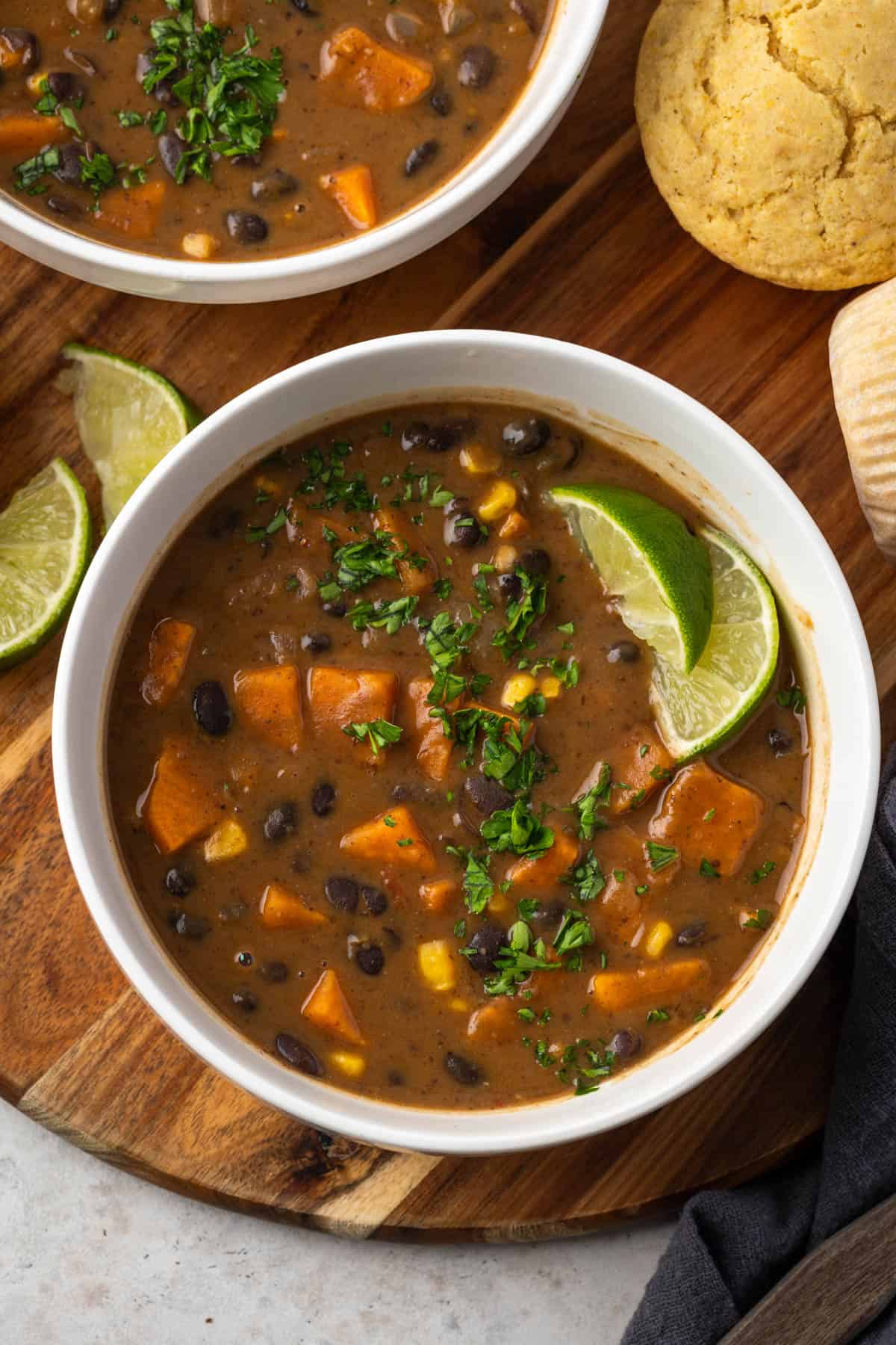 Two bowls of southwest black bean sweet potato soup on a wood platter nest to lime wedges and cornbread muffins.