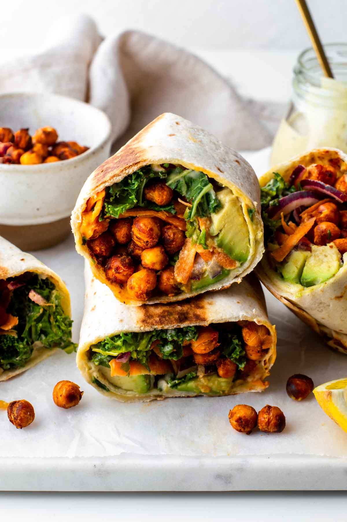 Kale and chickpea wraps on a table next to a bowl of roasted chickpeas and a jar of vegan caesar dressing.