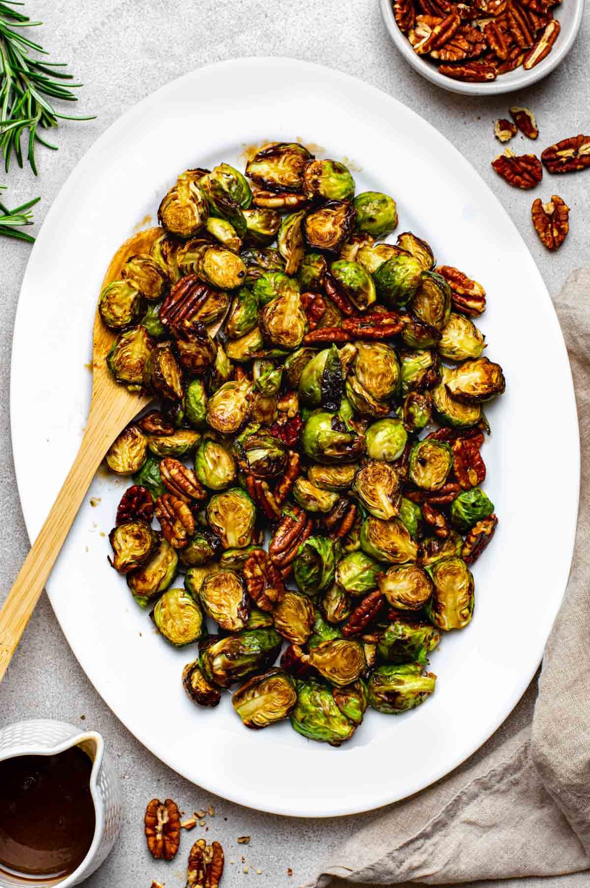 Air fryer maple balsamic brussels sprouts with pecans on a platter with a wooden spoon.