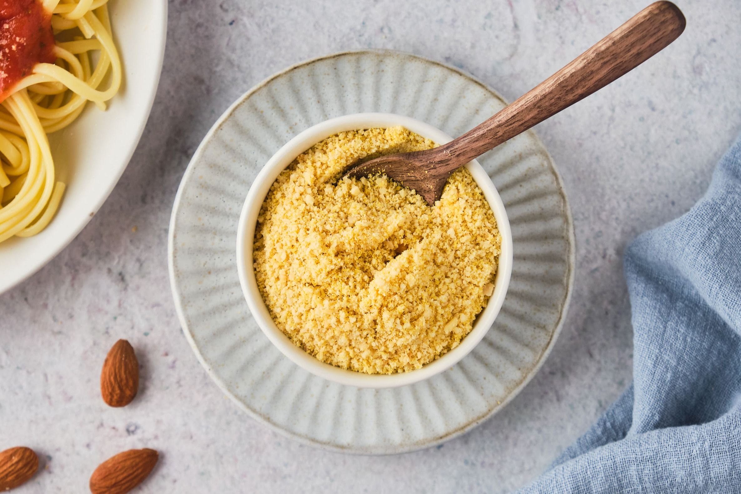 Vegan Parmesan cheese in a bowl with a wooden spoon next to a few almonds and a plate of spaghetti. 