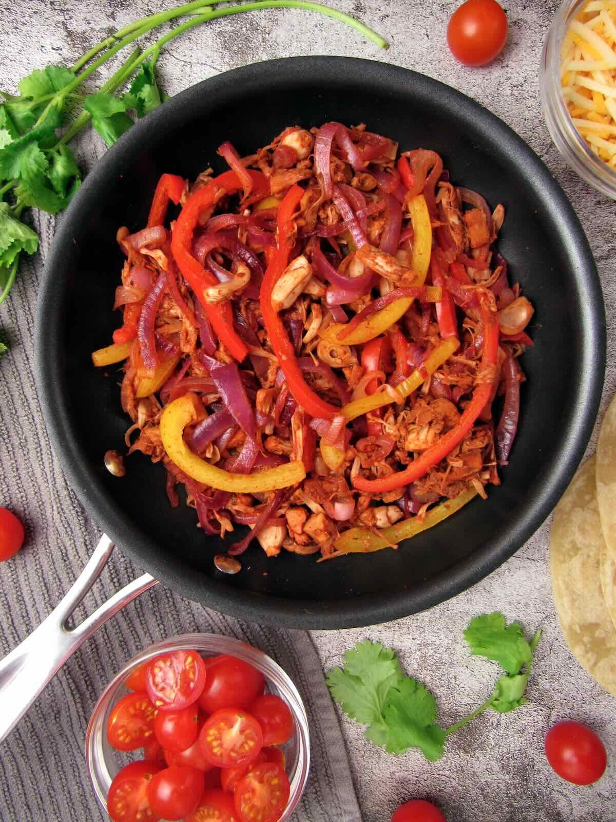 A pan of jackfruit veggie fajitas next to tortillas, cilantro, cherry tomatoes, and vegan cheese. 