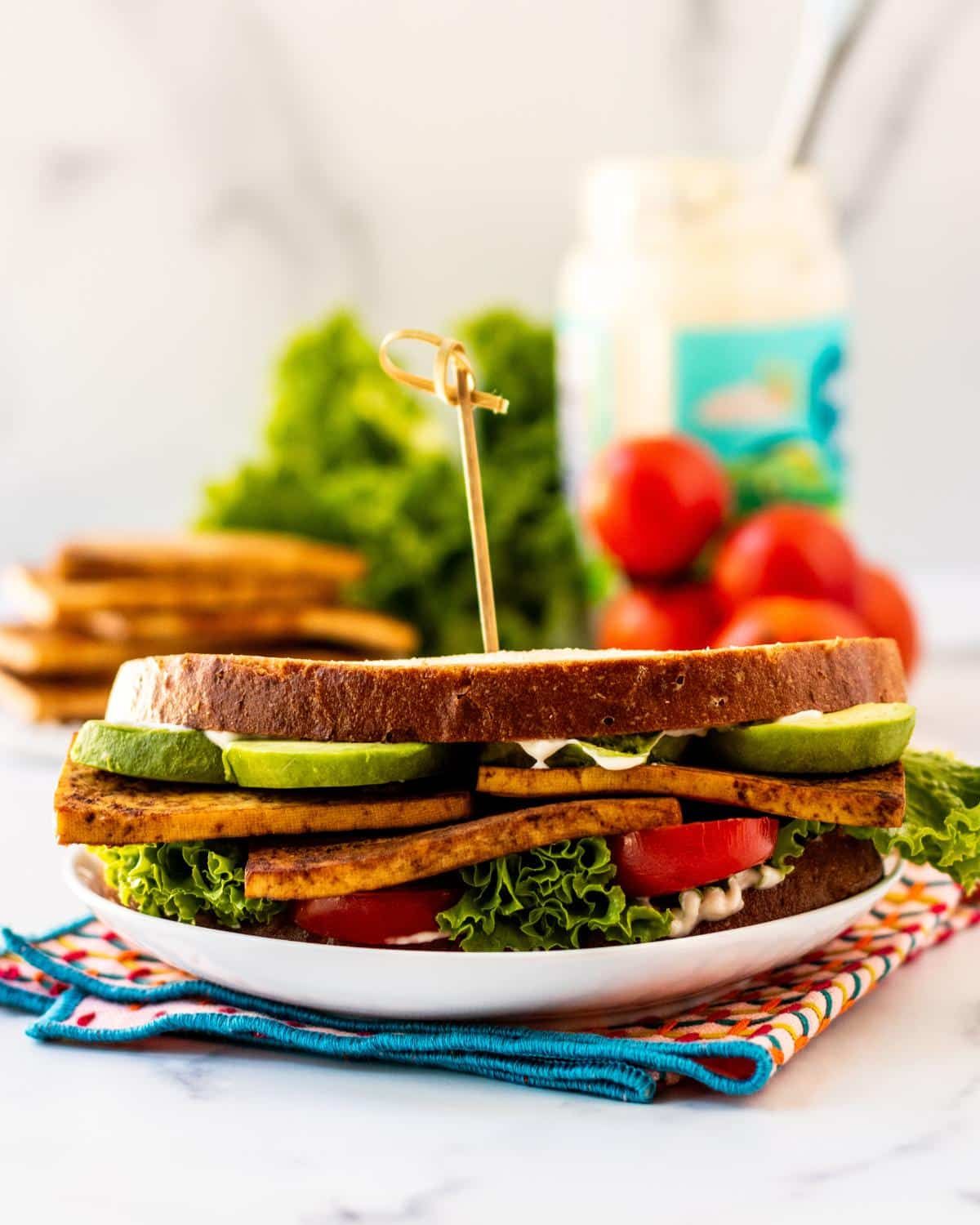 A baked tofu sandwich on a plate with a jar of vegan mayonnaise and baked tofu slices in the background. 