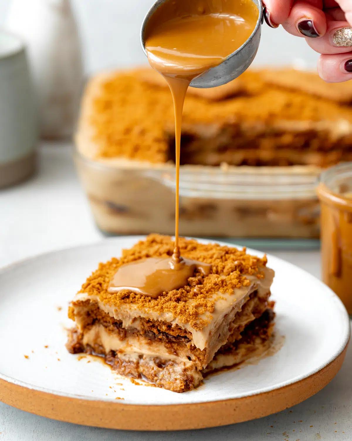 Sauce being poured over a slice of vegan Biscoff tiramisu, with the pan of tiramisu in the background.