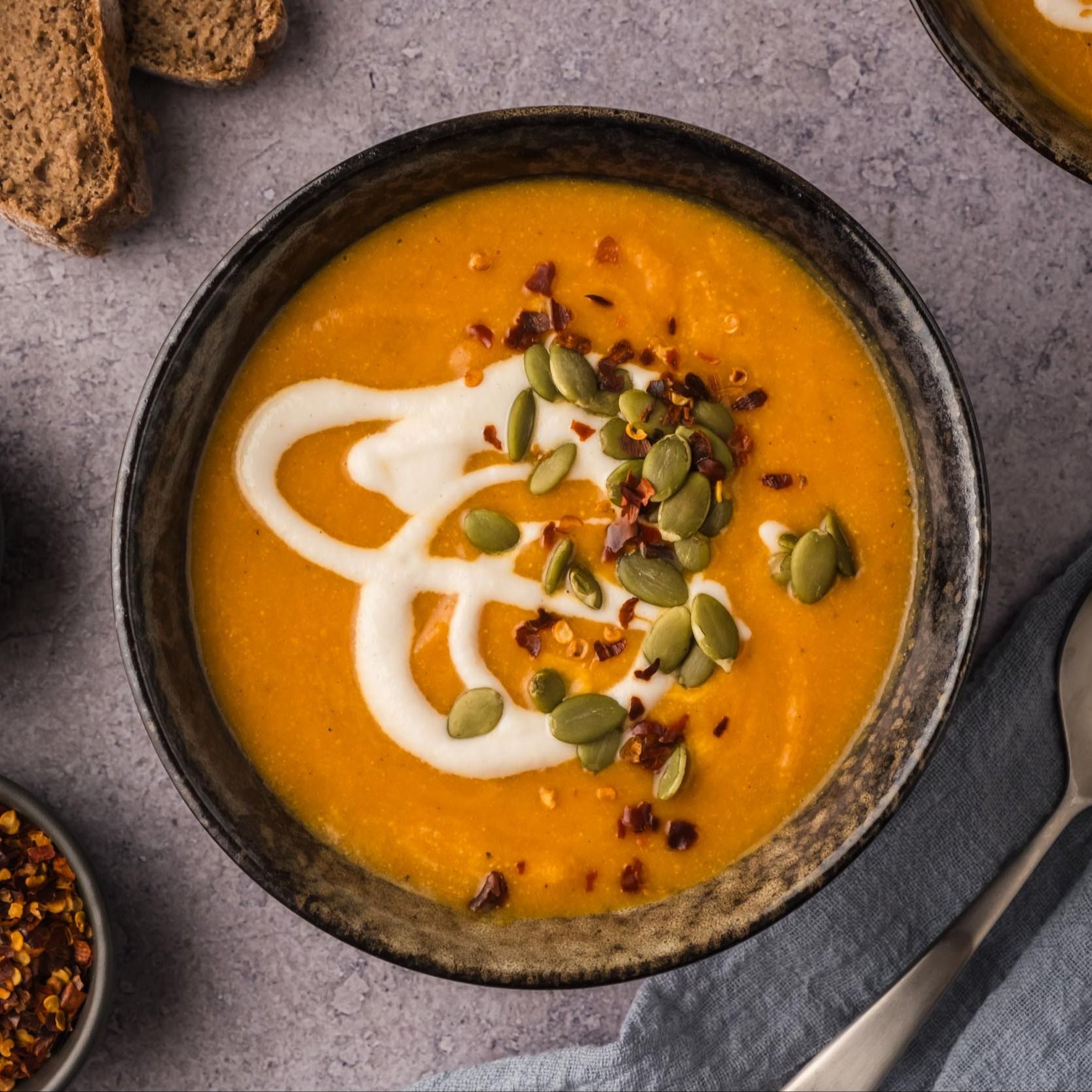 A top-down image of a bowl of creamy carrot pumpkin soup topped with cashew cream, red pepper flakes, and pepitas.