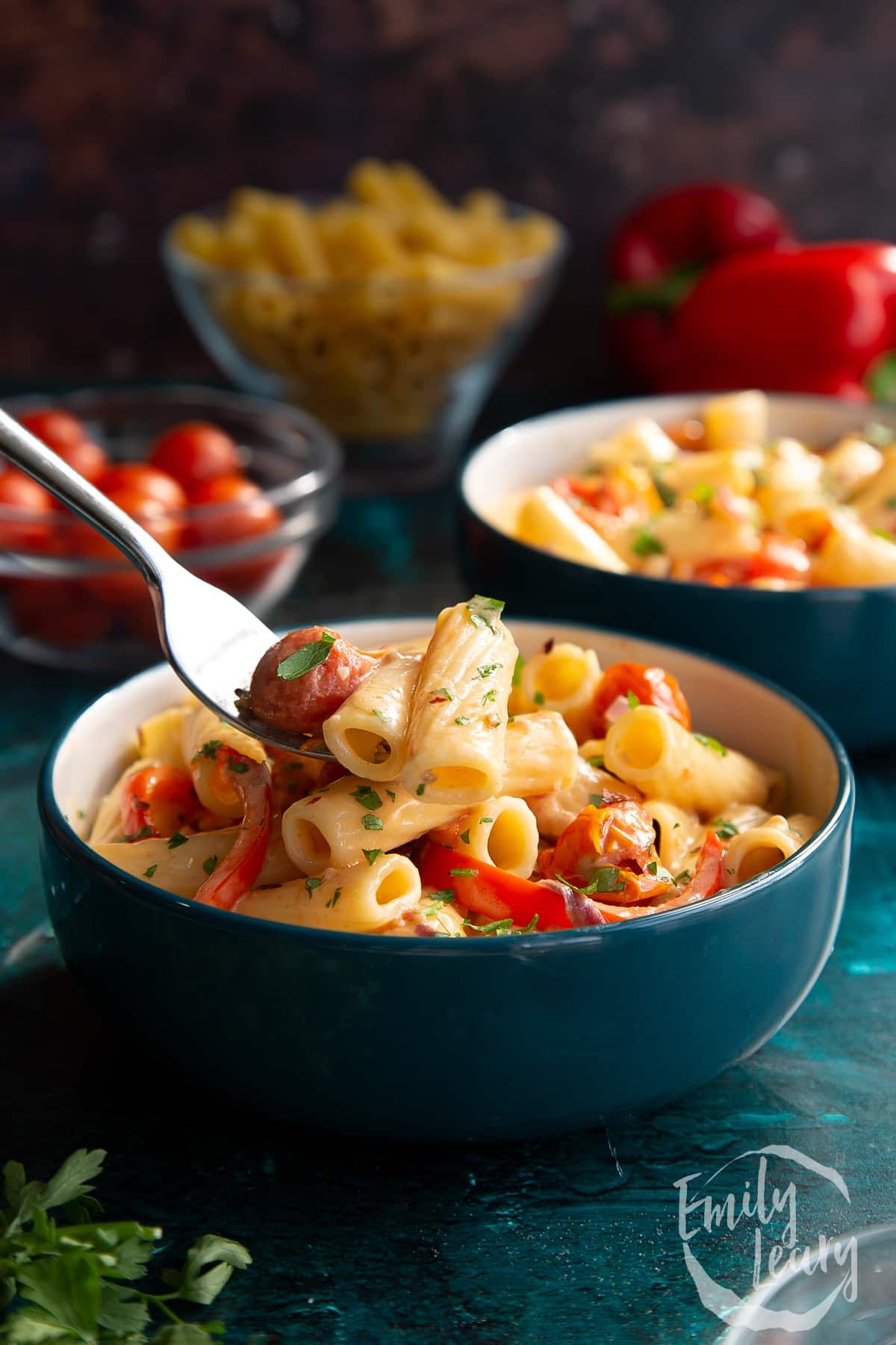 A fork taking some vegan feta pasta from a bowl, with red peppers, dry pasta, and cherry tomatoes in the background. 