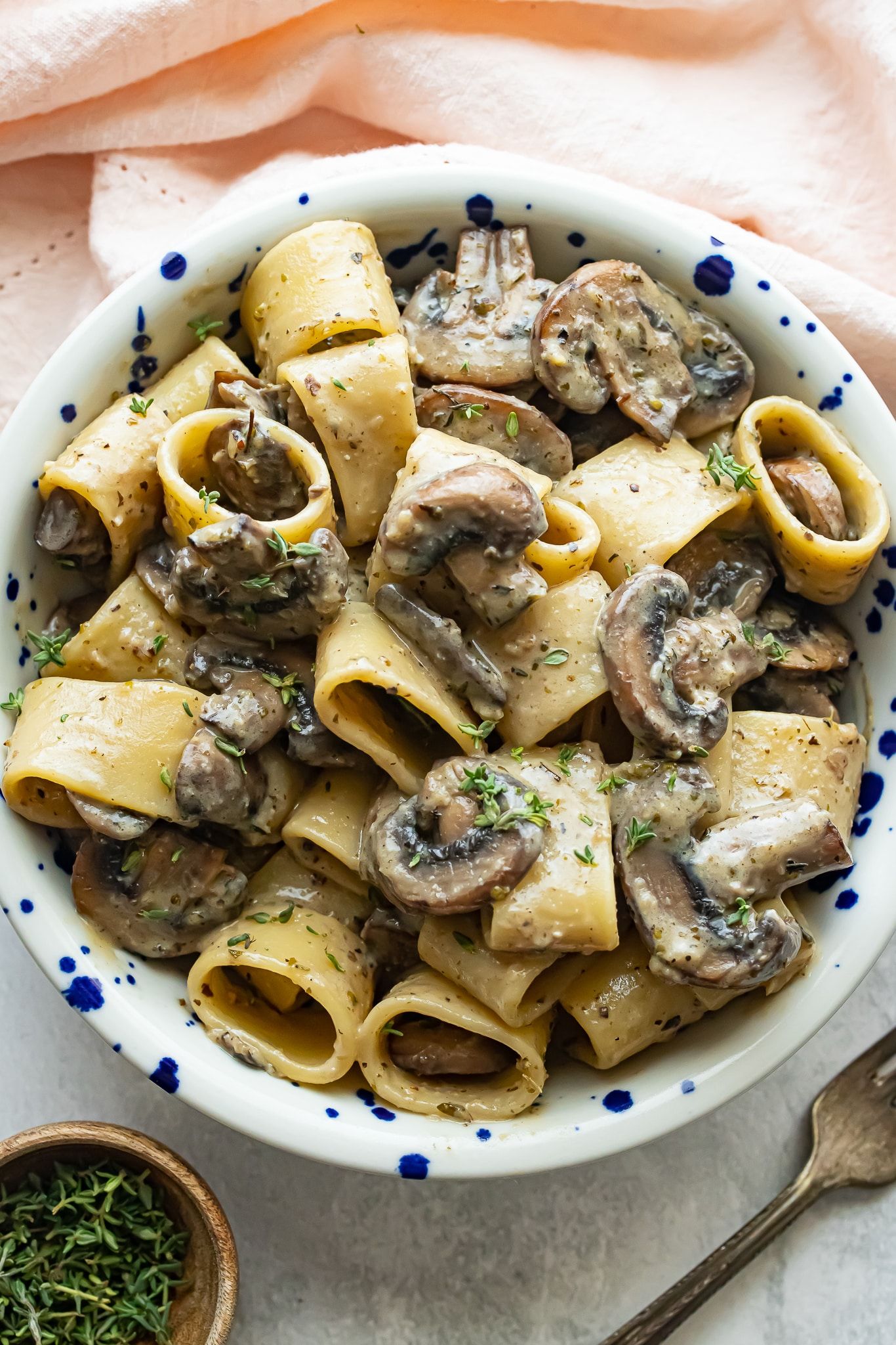 A bowl of miso mushroom pasta next to a fork and a pinch bowl of herbs. 