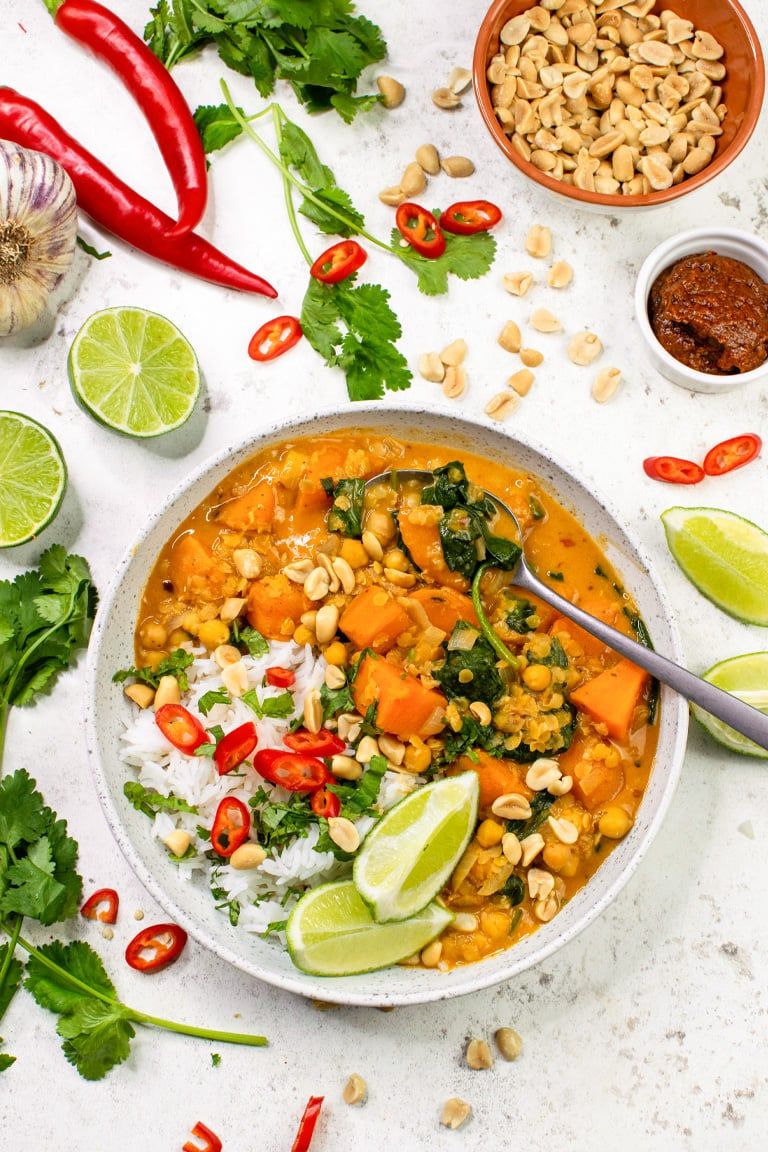 A table with various vegetables, limes, and peanuts, with a bowl of vegan red lentil curry. 