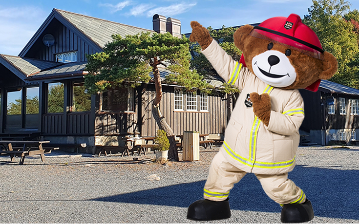 Stor bamse kledd i brannmannuniform står foran en trehytte
