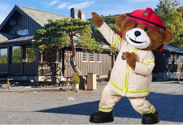Stor bamse kledd i brannmannuniform står foran en trehytte