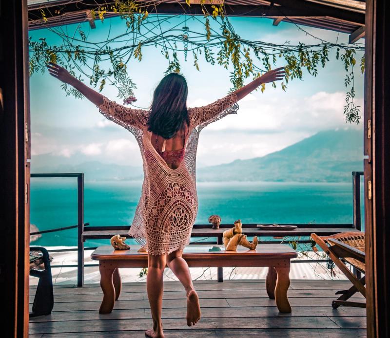 Frau mit ausgebreiteten Armen, die von der Terrasse ihres Zimmers aus auf einen türkisfarbenen See blickt