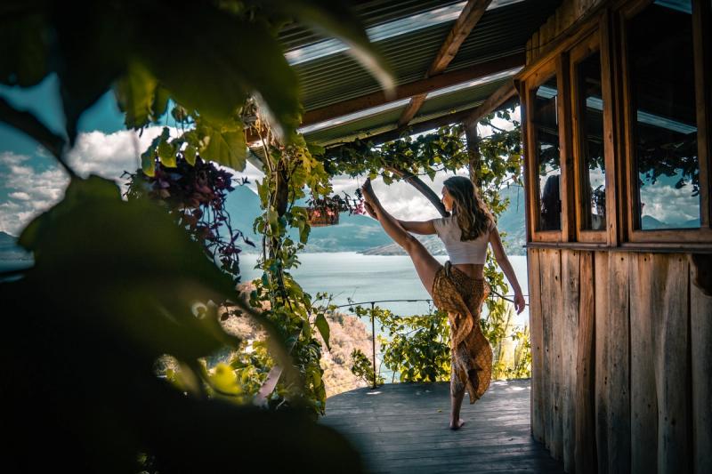 Frau, die auf einer Terrasse vor dem Hintergrund eines Vulkans und eines Sees eine Yoga-Pose aus dem „Dancer’s Yoga“ ausführt